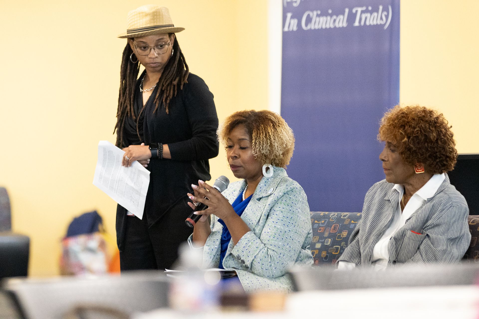 Three women speaking at a table. One in a hat stands behind them, holding papers. Blue banner in background.