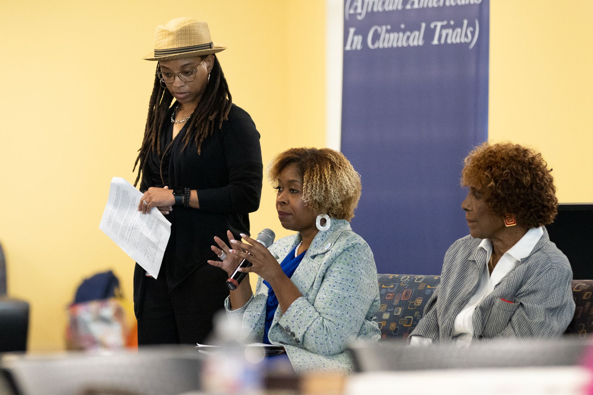 Panel of women speaking at event. One holds a microphone, another reads, and the third listens. Banner in background.