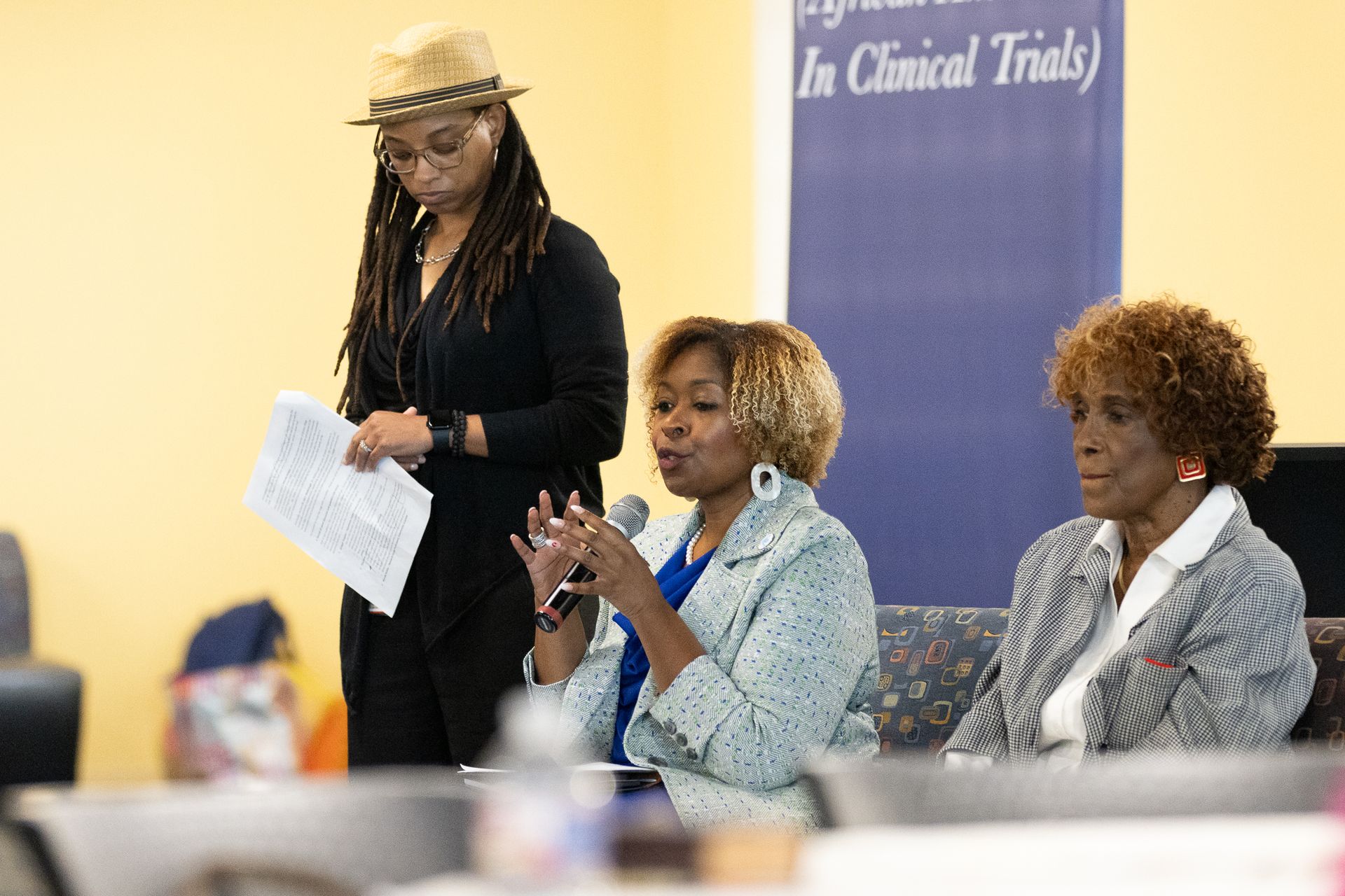 Panel discussion with three women seated, one speaking into a microphone. Another woman stands holding papers.