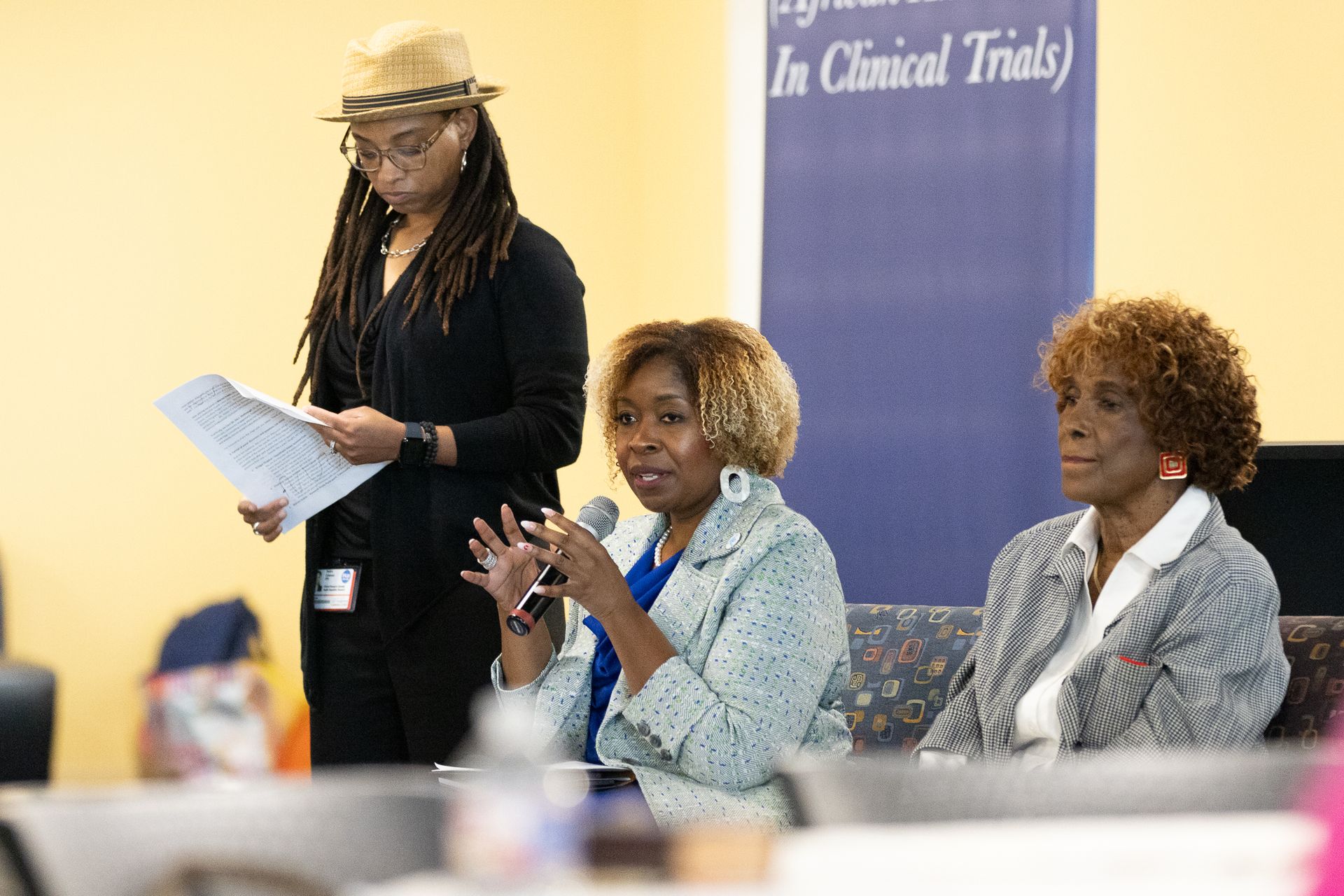 Panel discussion: Three women seated, speaking into microphones, and a woman standing, holding papers.