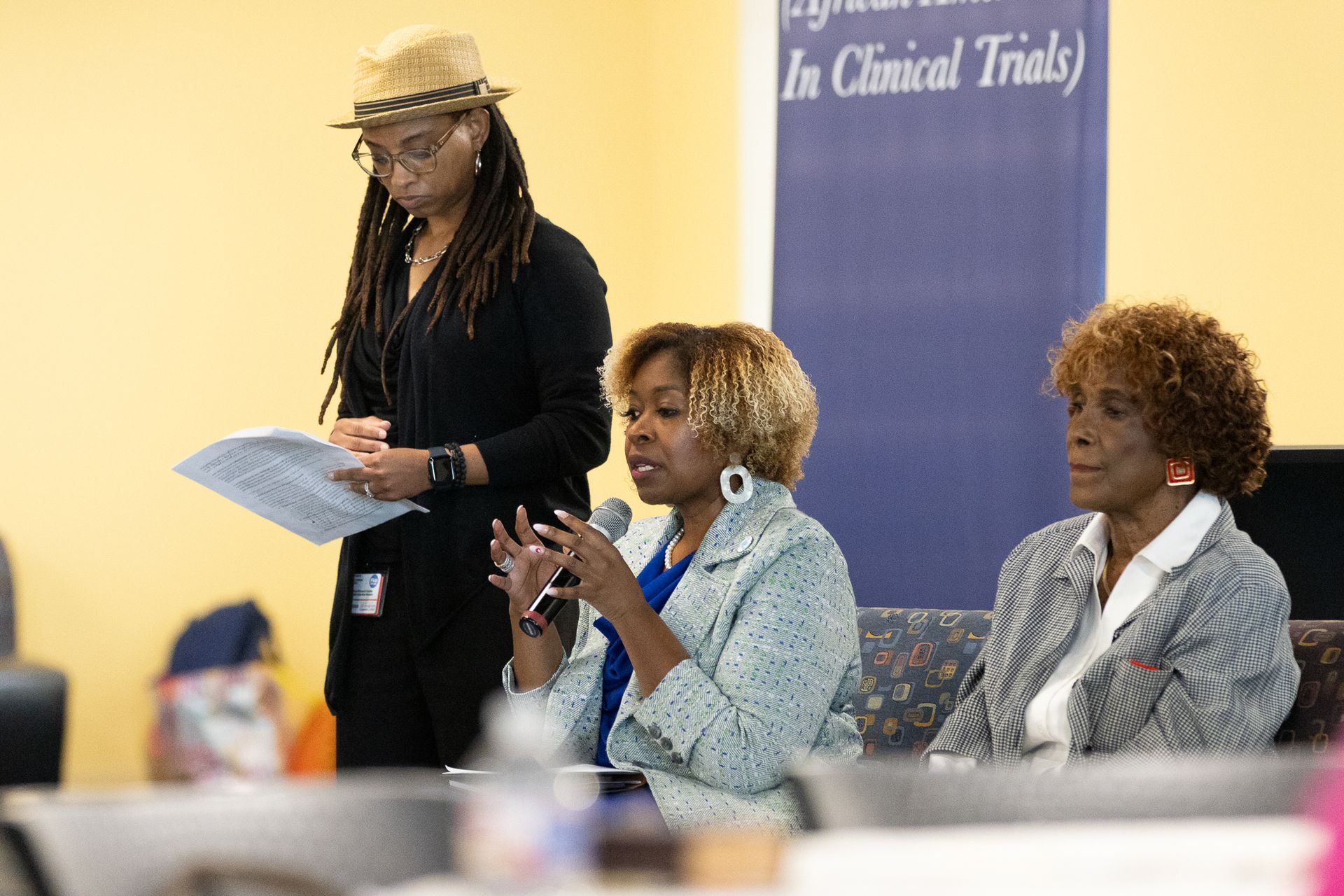 Three women at a table; one speaks into a microphone, another looks on. A fourth person, standing, holds papers.
