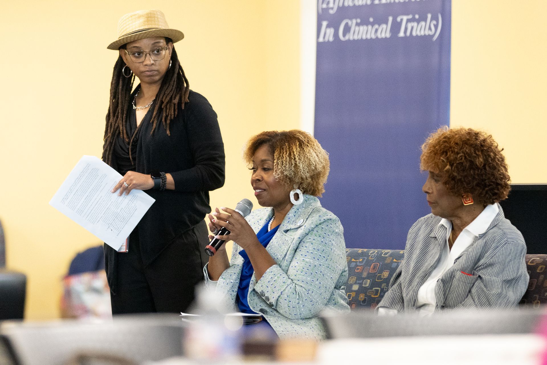 Panel of three women; one speaks into a microphone, another holds papers, and the third sits and listens.