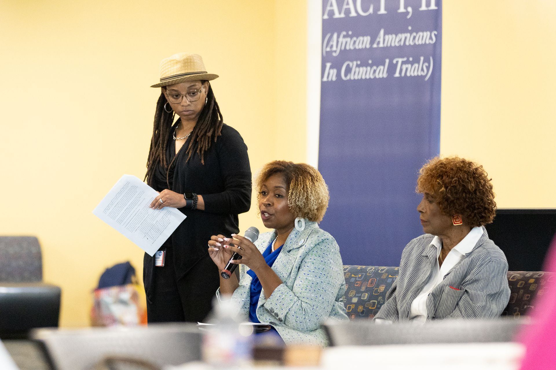 Panel of women speaking at an event, with banner reading “African Americans In Clinical Trials.”