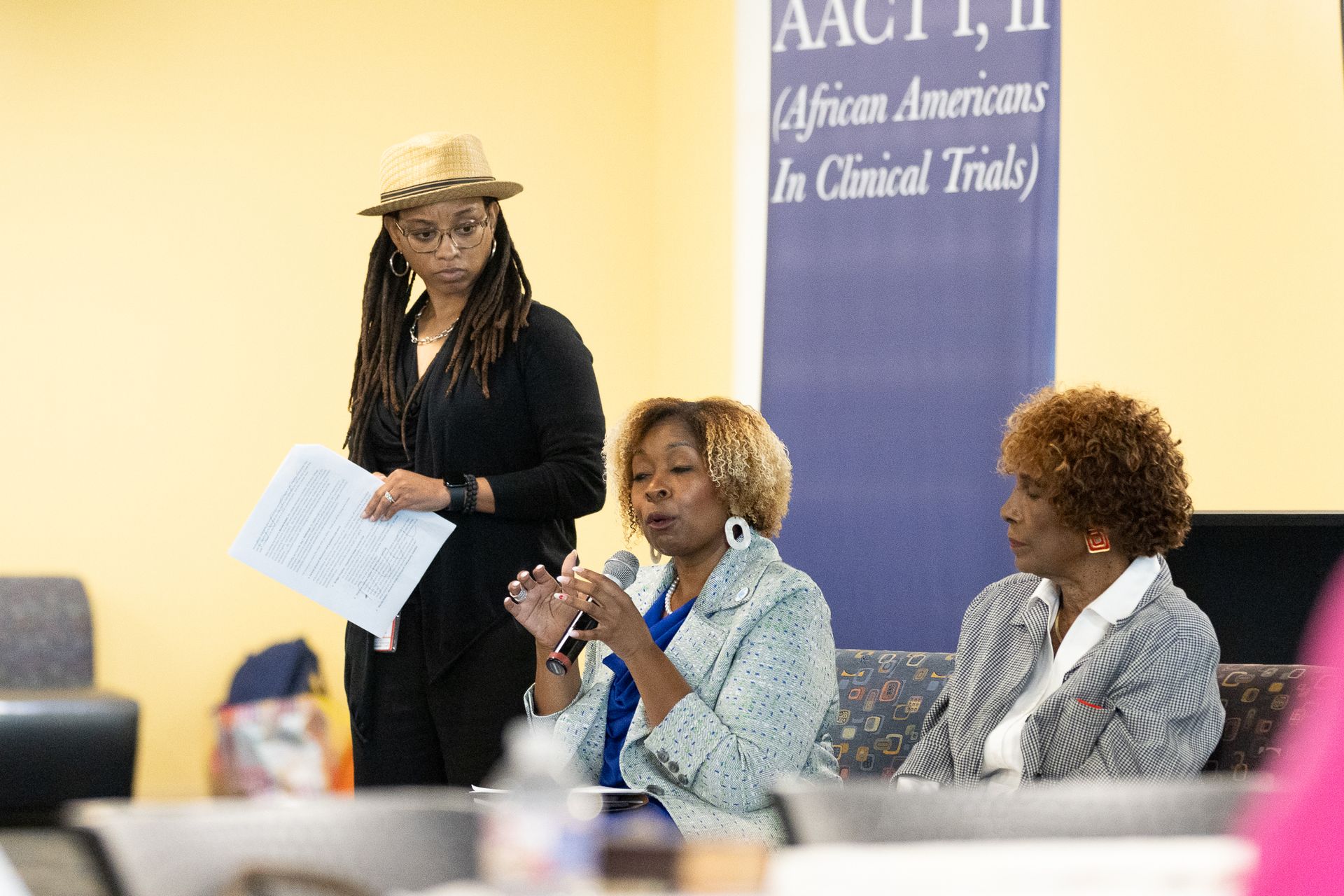 Panel discussion with three women at a conference, one speaking into a microphone, another holding papers.