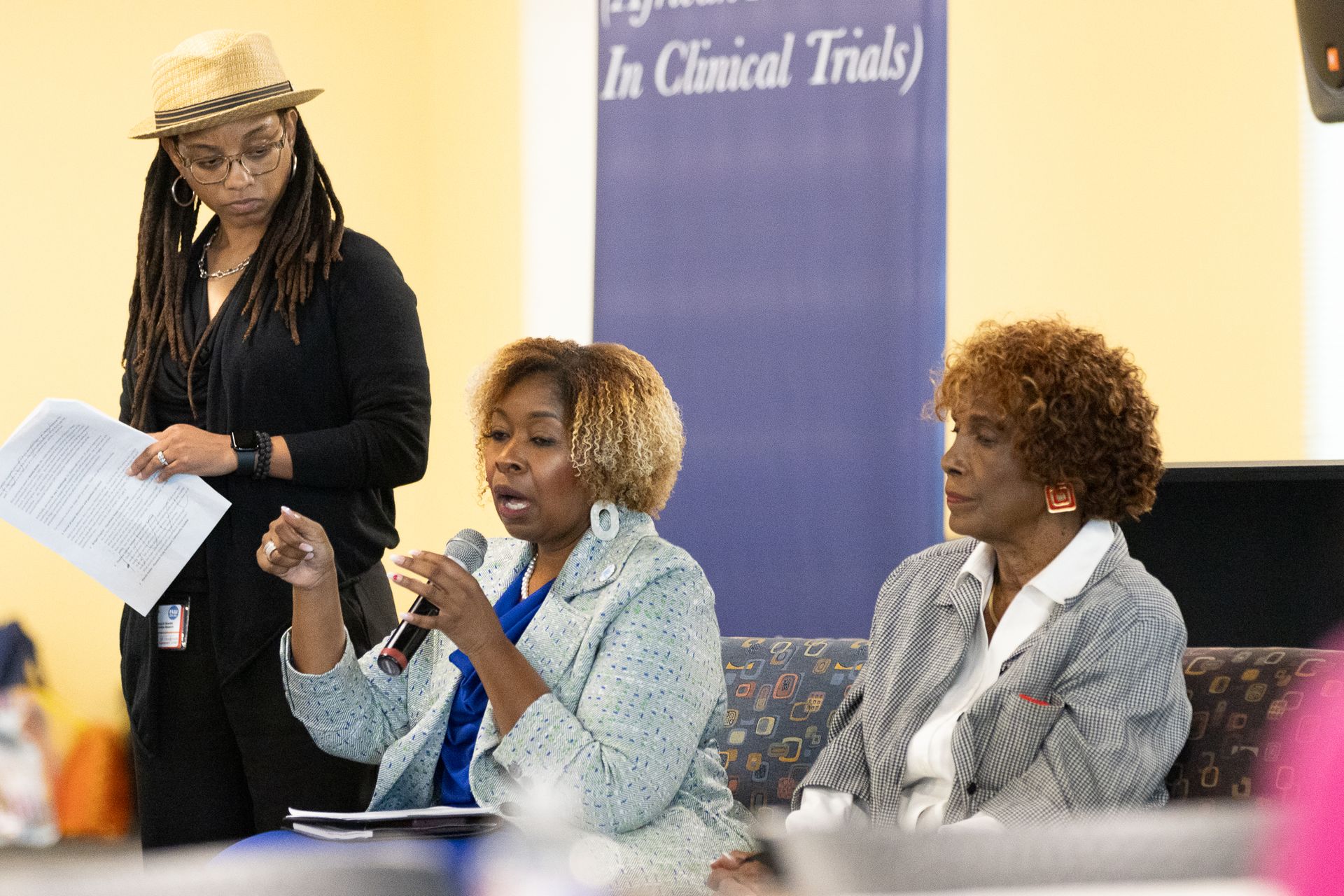 Three women at a panel discussion; one speaking into a microphone. Banner in the background.