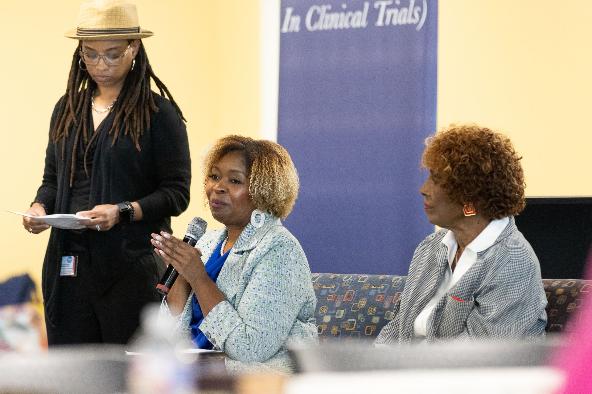 Three people at an event: two women seated, one speaking with a microphone; another standing to the side holding papers.
