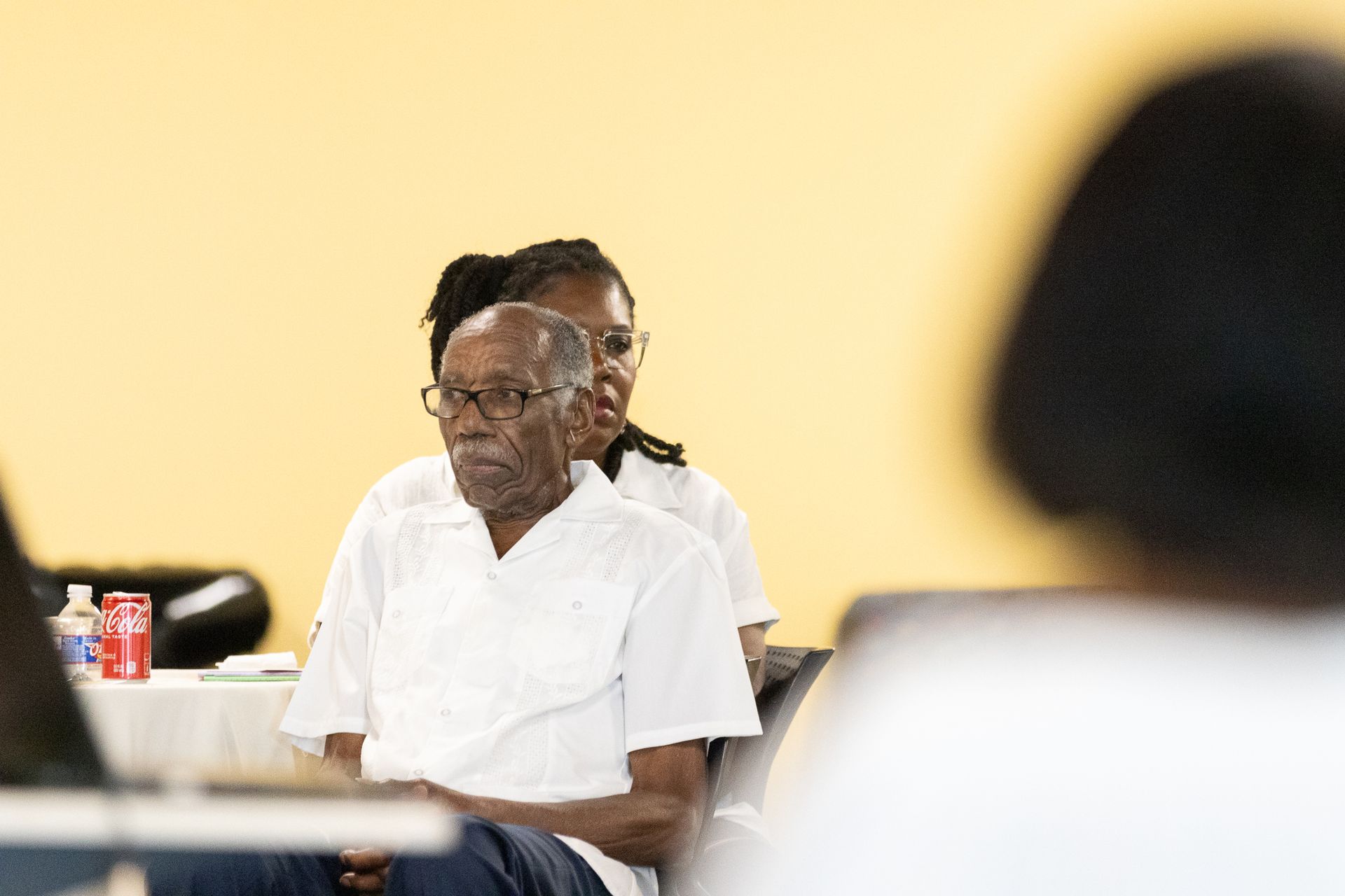 Man in white shirt, wearing glasses, seated, listening in a brightly lit room.