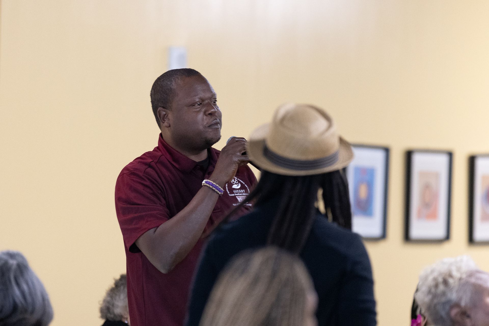 Man speaking in a room; another person wearing a fedora in the foreground. Cream walls and framed art in the background.