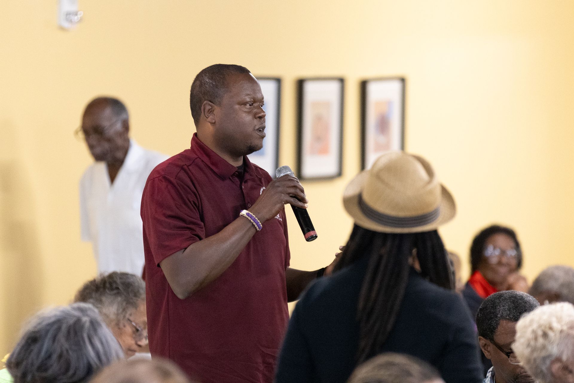 Man in maroon shirt speaks into a microphone at a gathering. Other people seated around him.