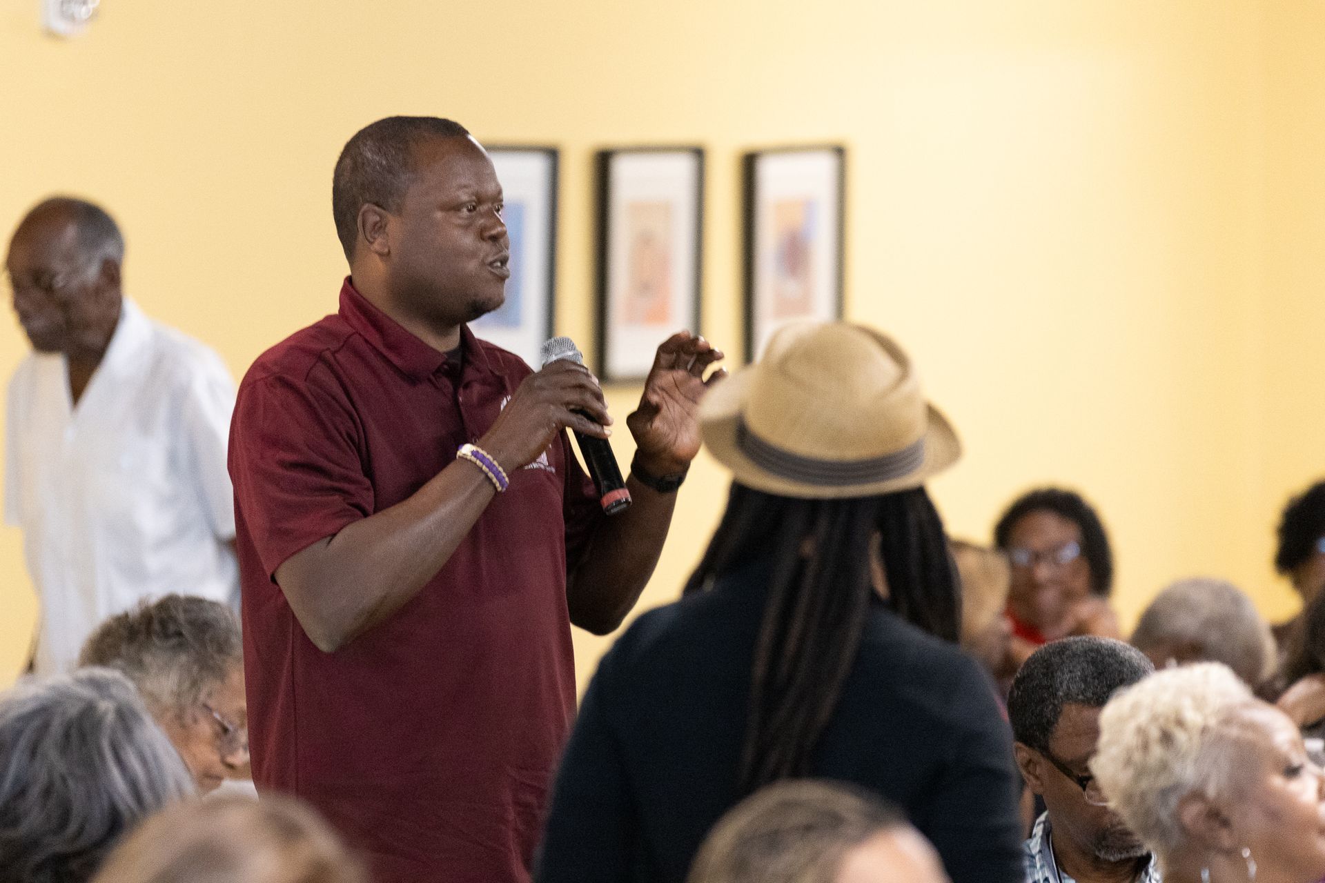 Man speaking into a microphone, addressing a crowd in a room with paintings on the wall.