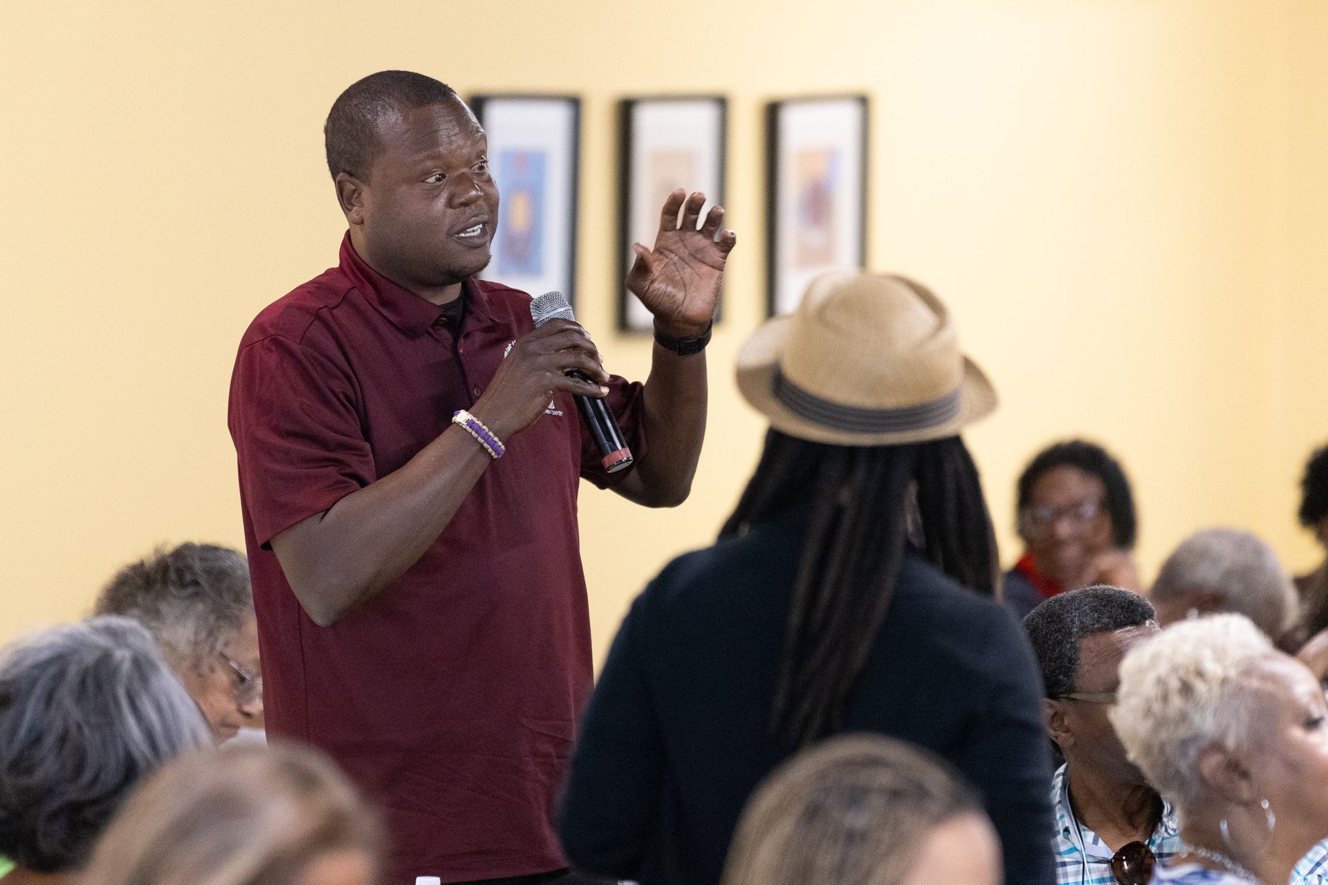 Man in maroon shirt speaks into a microphone at a gathering. Other people seated, listening.