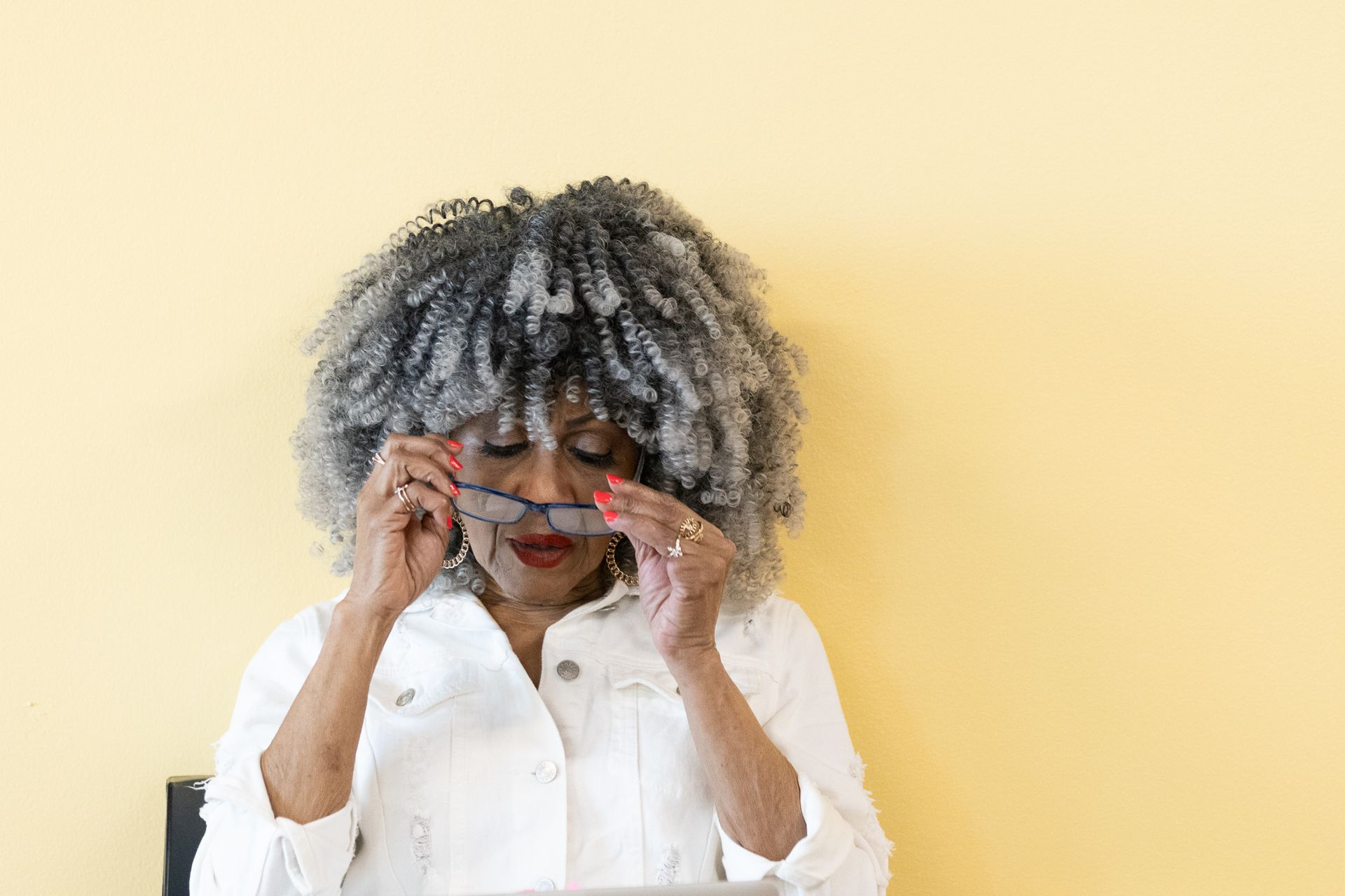 Woman in white jacket and gray hair adjusts glasses in front of a yellow wall.