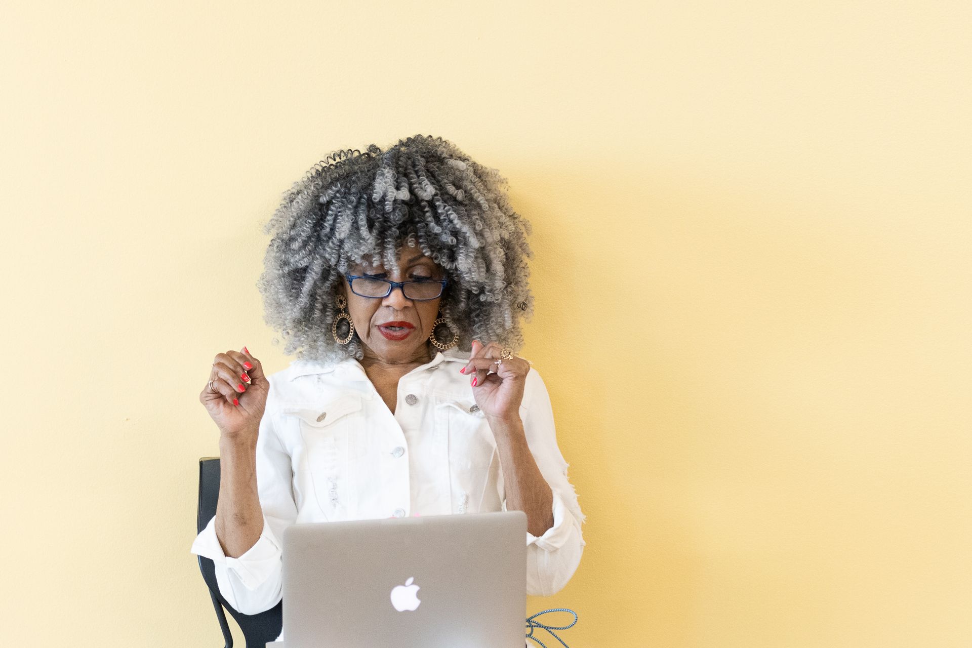 Woman with gray hair and glasses looks at laptop, arms raised, sitting against a yellow wall.