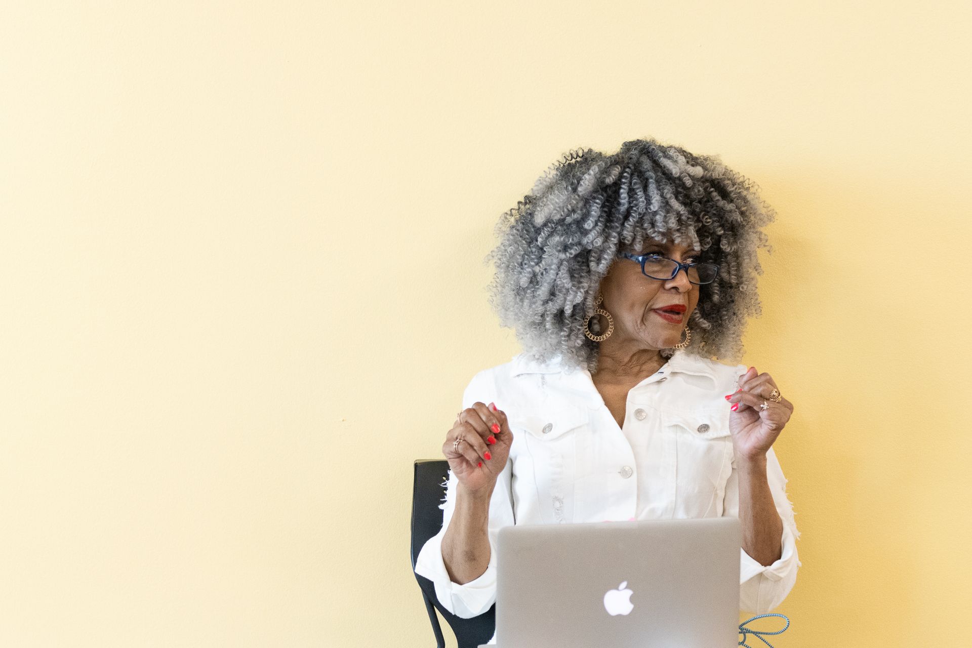 Woman with gray, curly hair sits at a laptop, looking right, with hands raised. Bright yellow background.