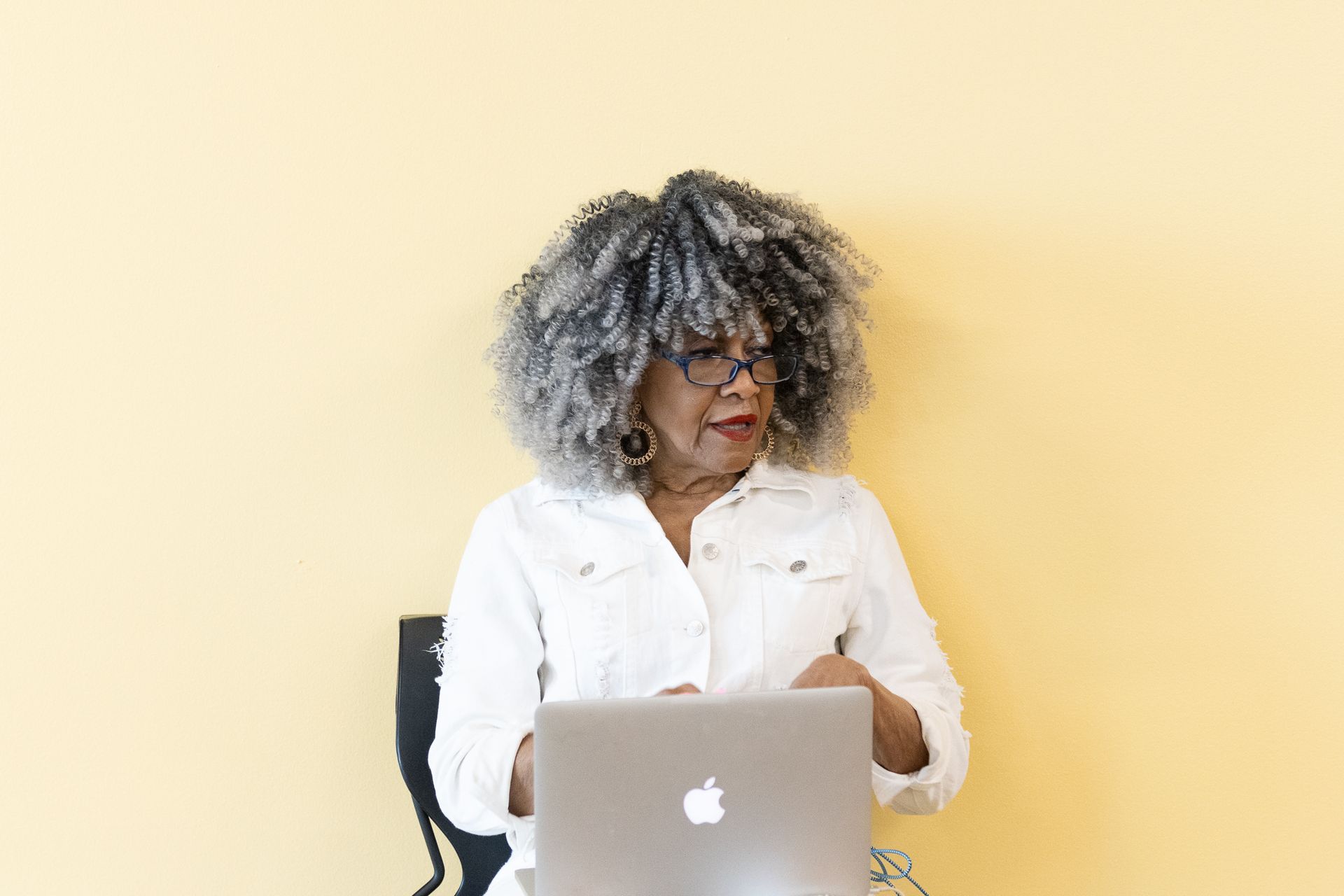 Woman with grey, curly hair looking right, using laptop, white jacket, yellow wall.