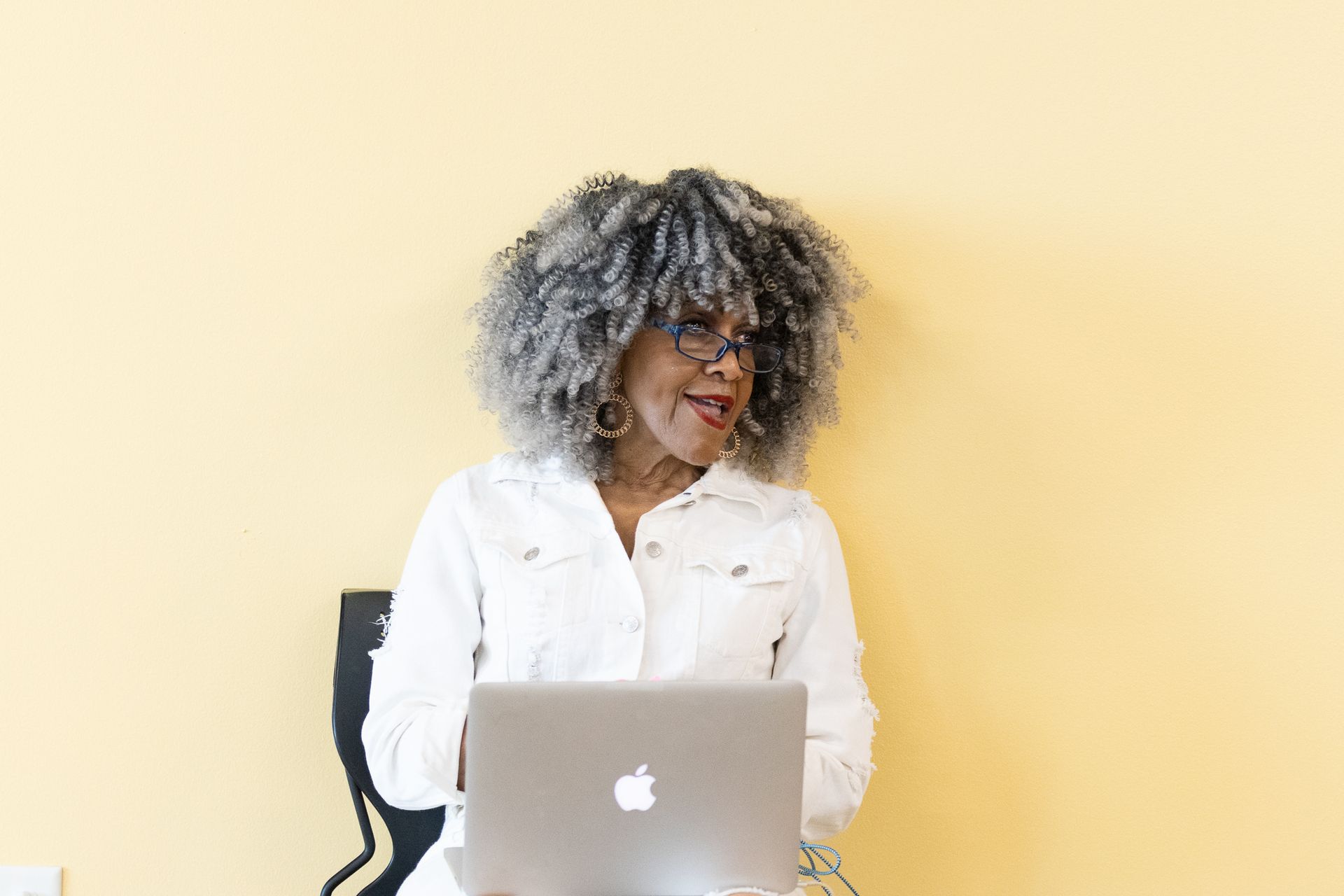 Woman with gray afro, in white denim jacket, using laptop, leaning against yellow wall.