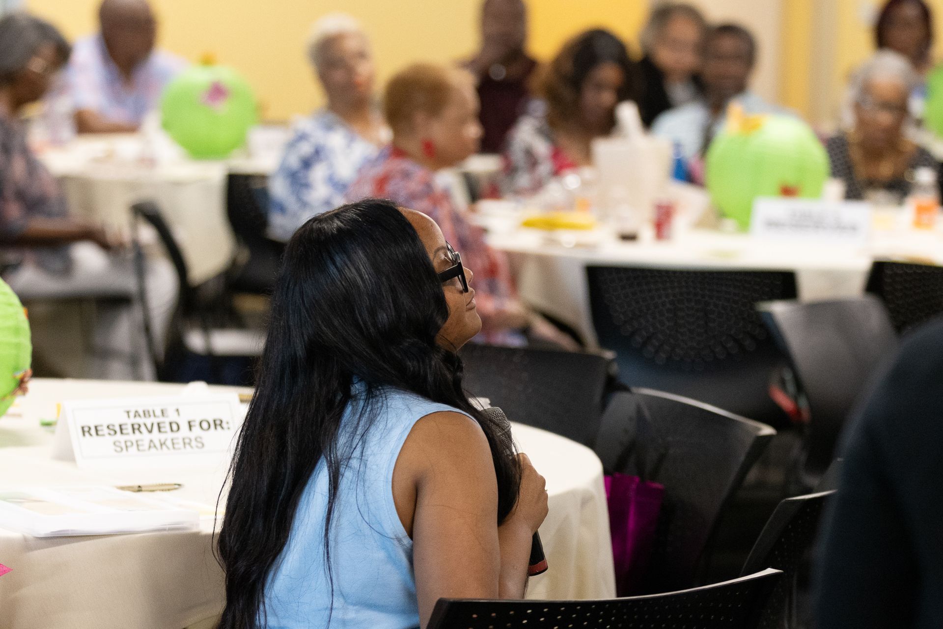 Woman speaking at event; tables with attendees, green decorations. 