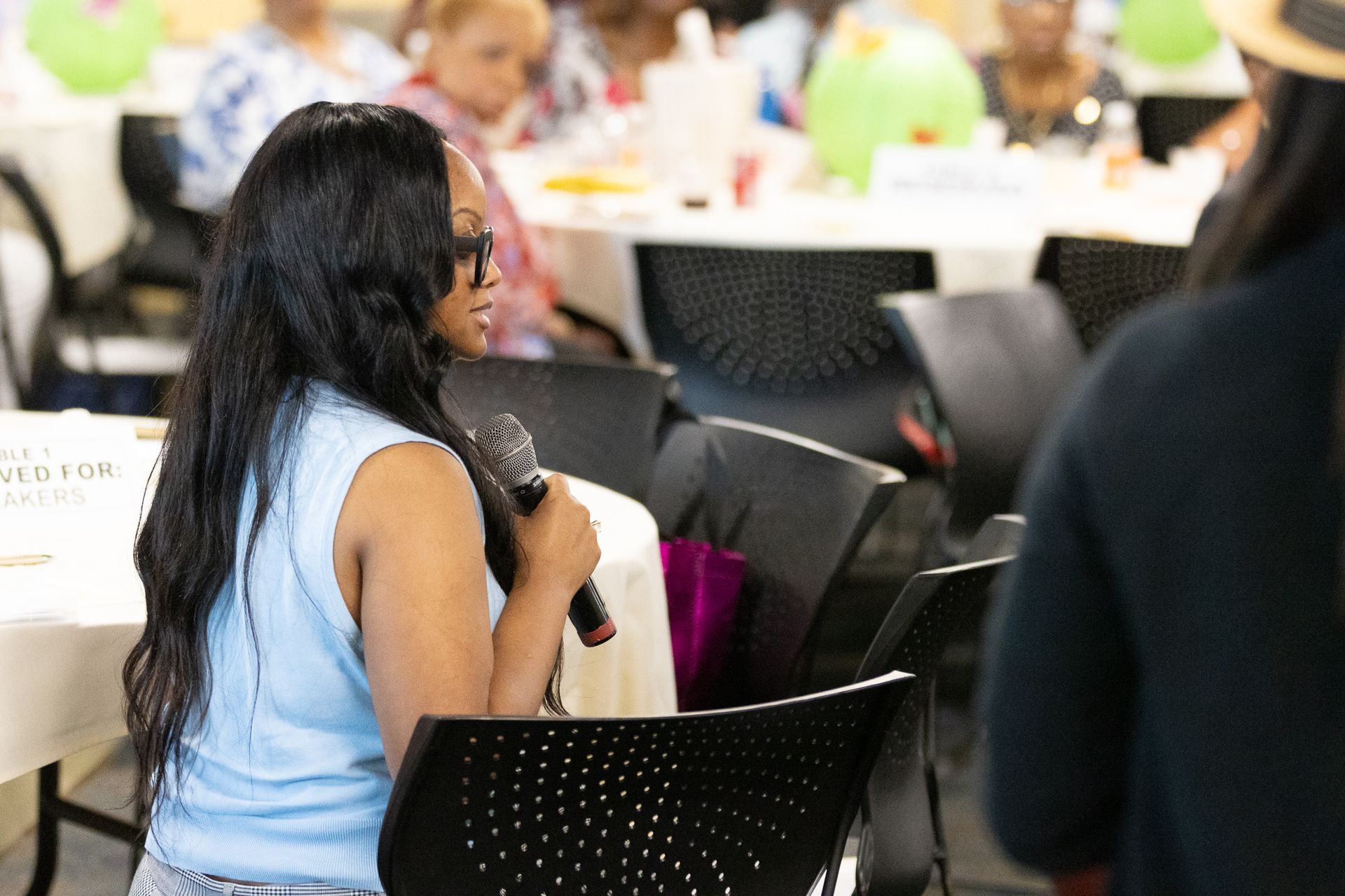 Woman with long dark hair speaks into a microphone at a round table; others in the background.