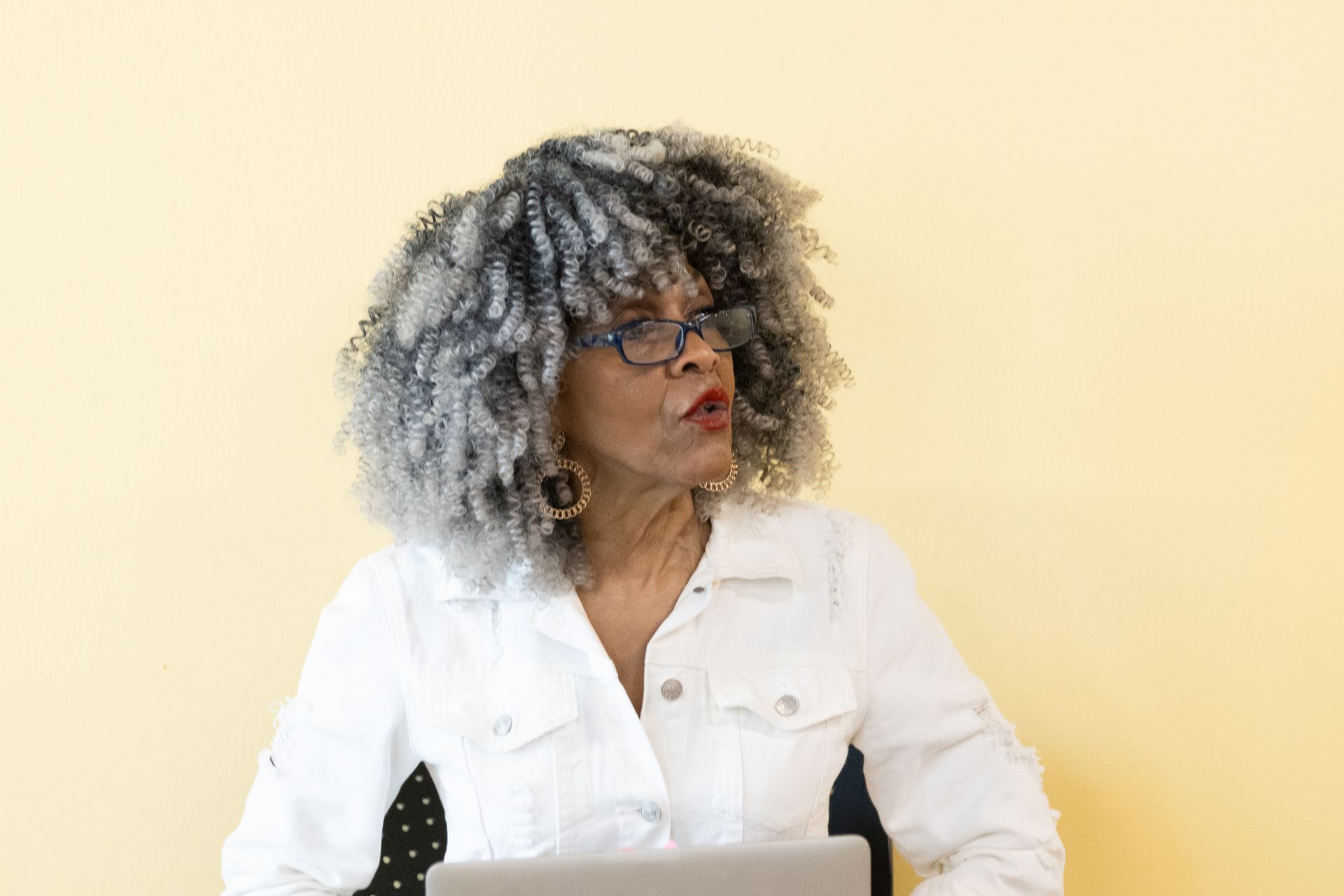 Woman with gray curly hair, wearing glasses and white jacket, looking to the side, near a laptop.