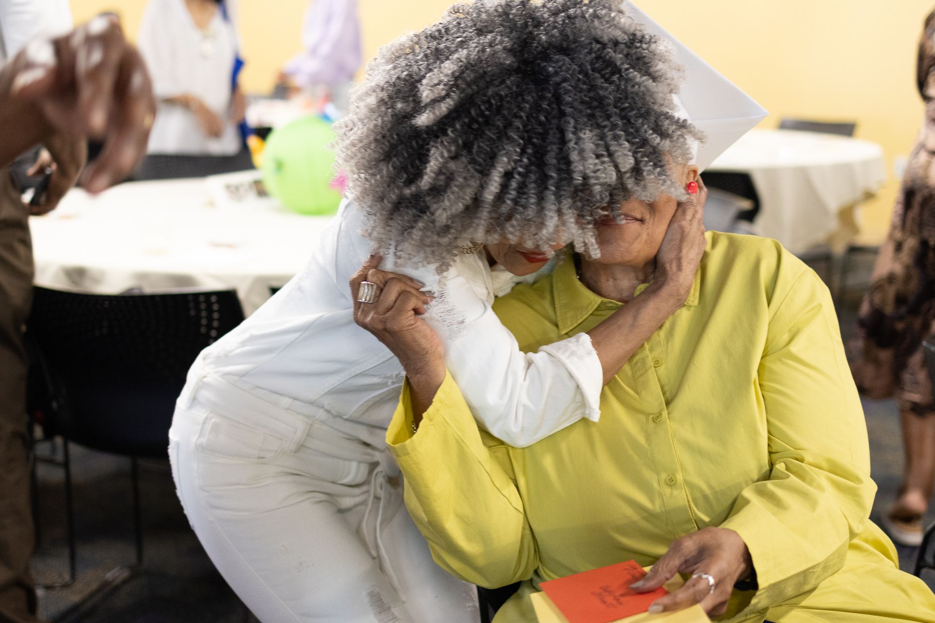 Two women embrace, one with gray afro, other in yellow. Room with tables and people.