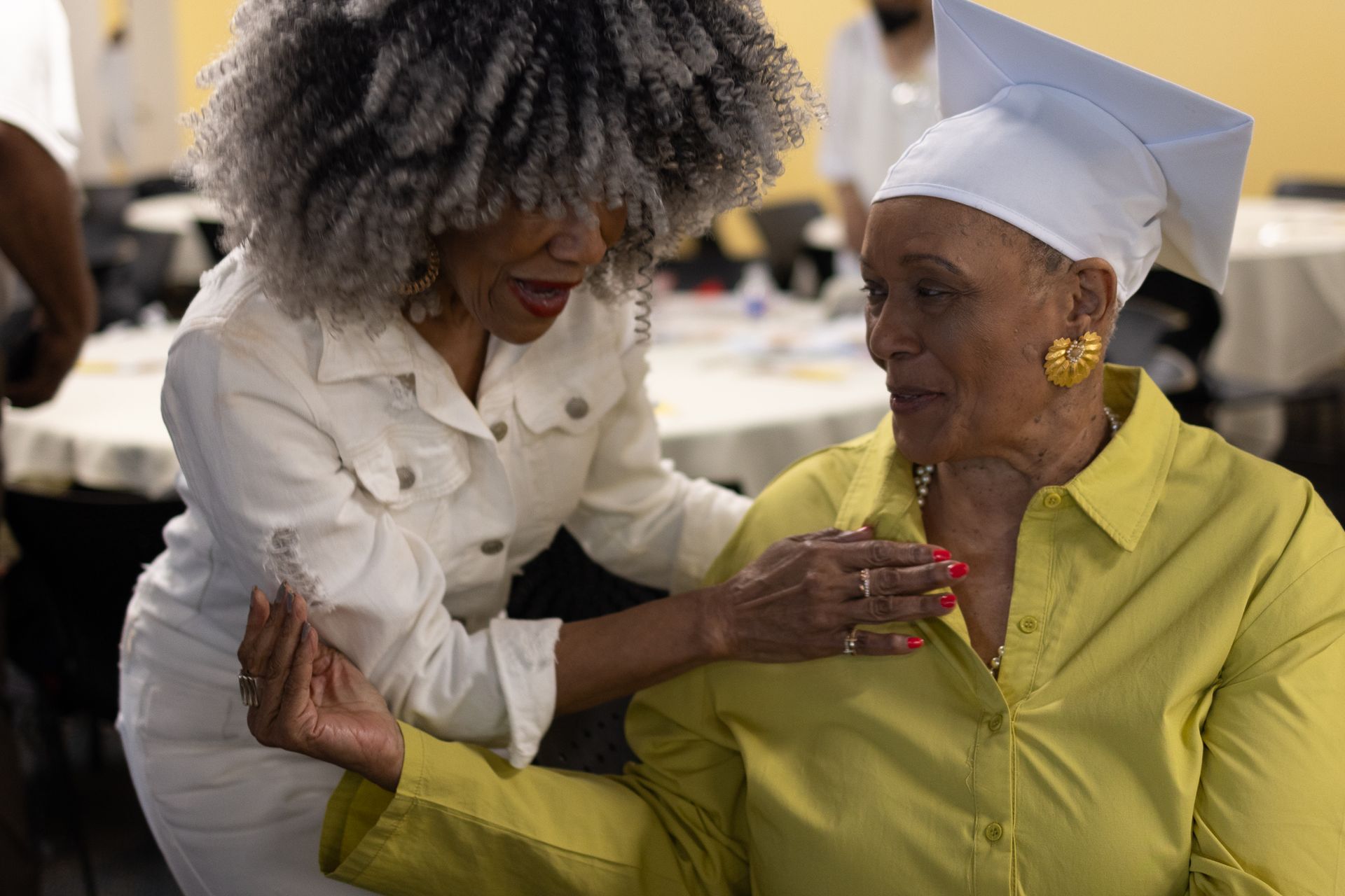 Woman in white jacket adjusting the graduation cap of another woman in a yellow shirt.