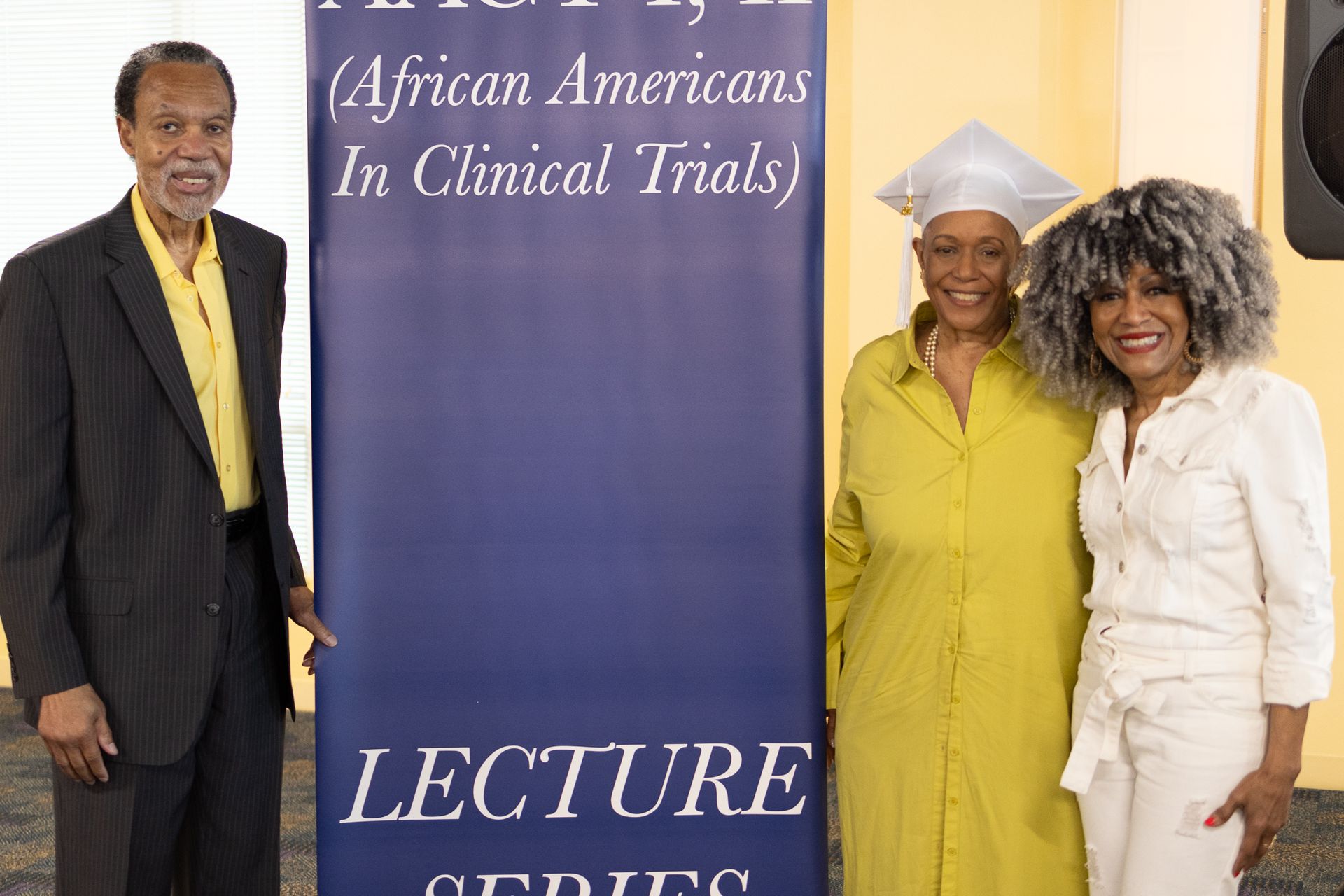 Three people pose by a blue banner that says “African Americans In Clinical Trials” during a lecture.
