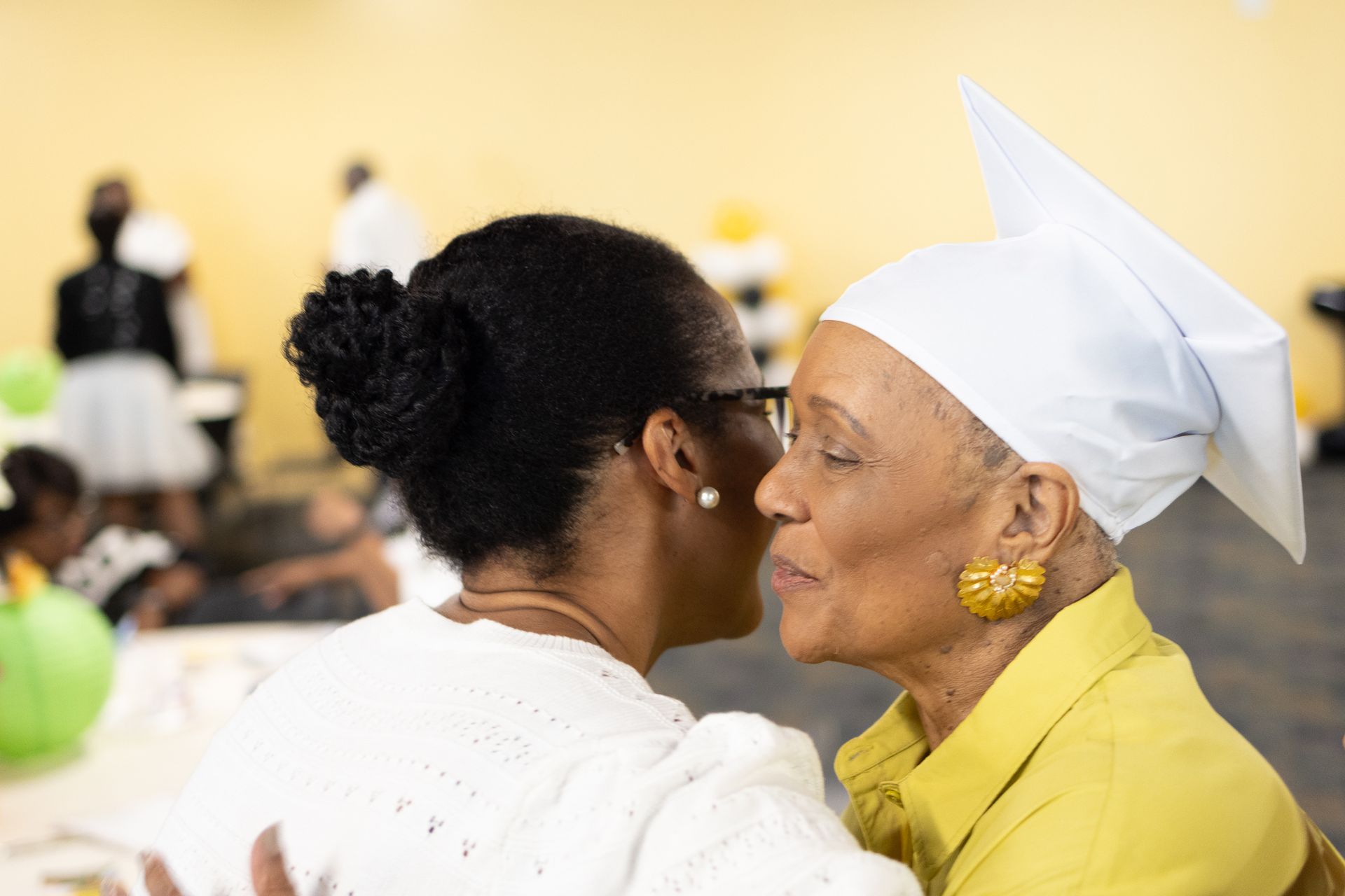 Woman in a graduation cap kissed by another woman. Celebration with yellow and white decorations.