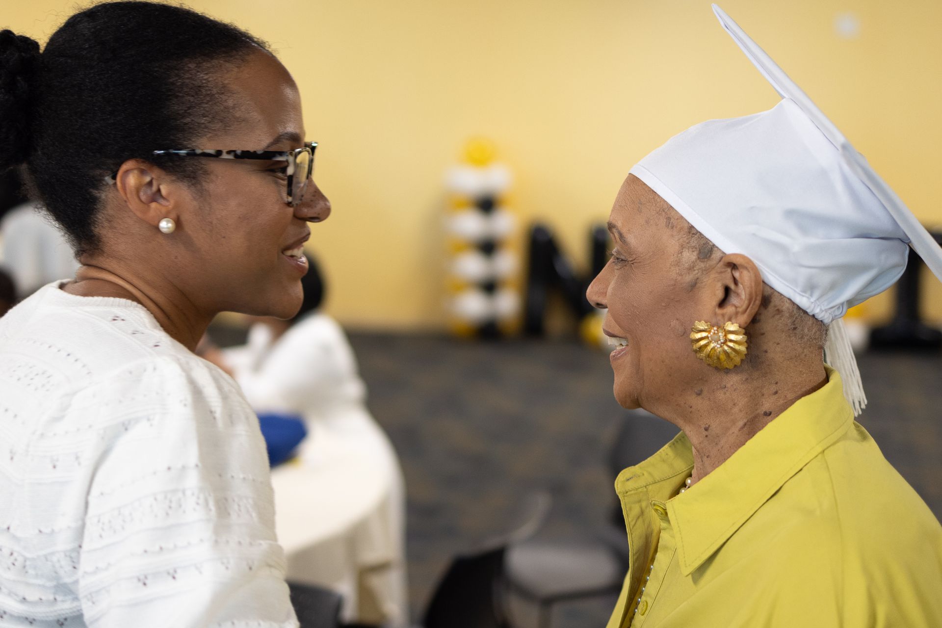 Woman in glasses talks to woman in cap and yellow jacket, both smiling, inside a room.
