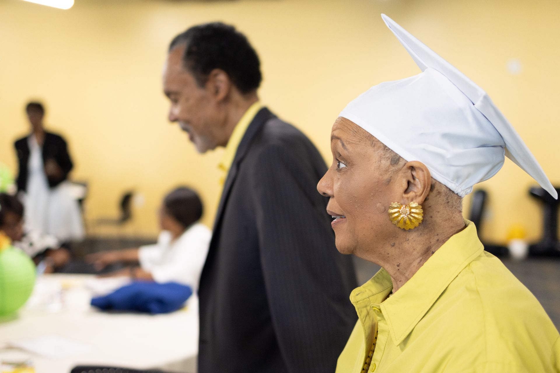 Man and woman in a room, the woman wearing a graduation cap. The woman is wearing a yellow shirt and large earrings.