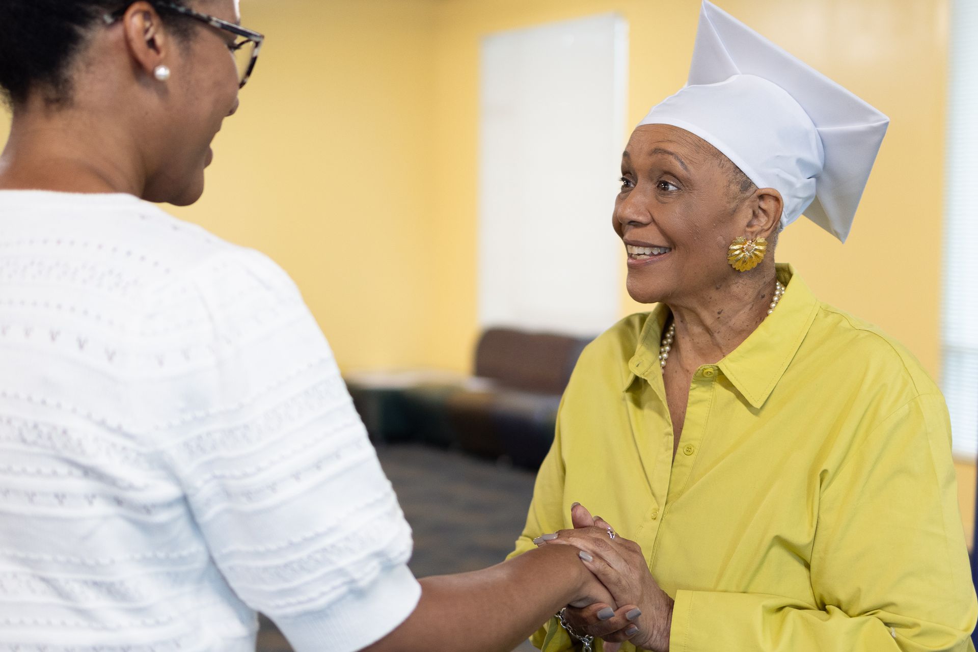 Woman in cap, yellow shirt, shakes hands with another woman, both smiling in a room with yellow walls.