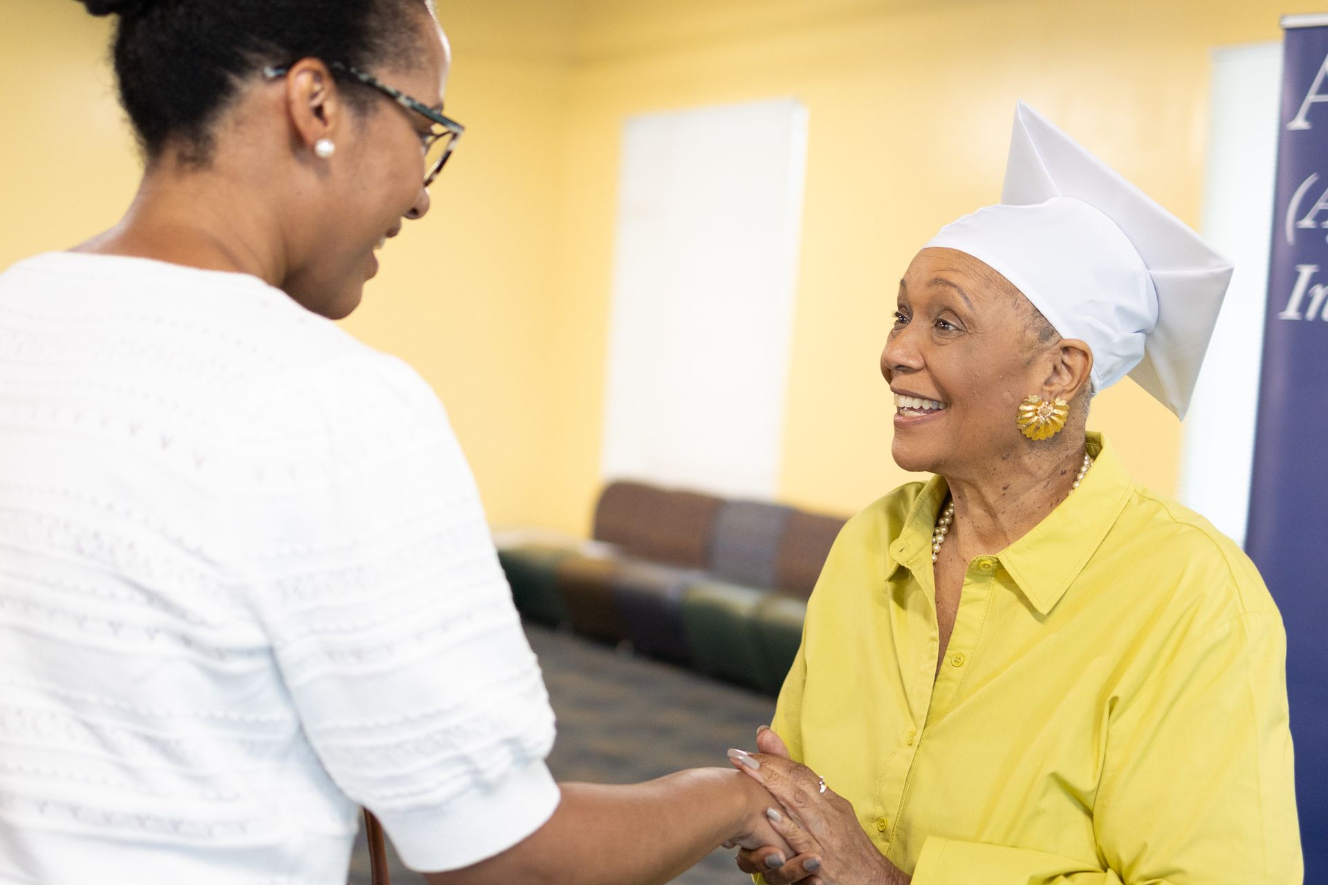 Woman in graduation cap smiles at another woman, holding her hand; indoor setting.