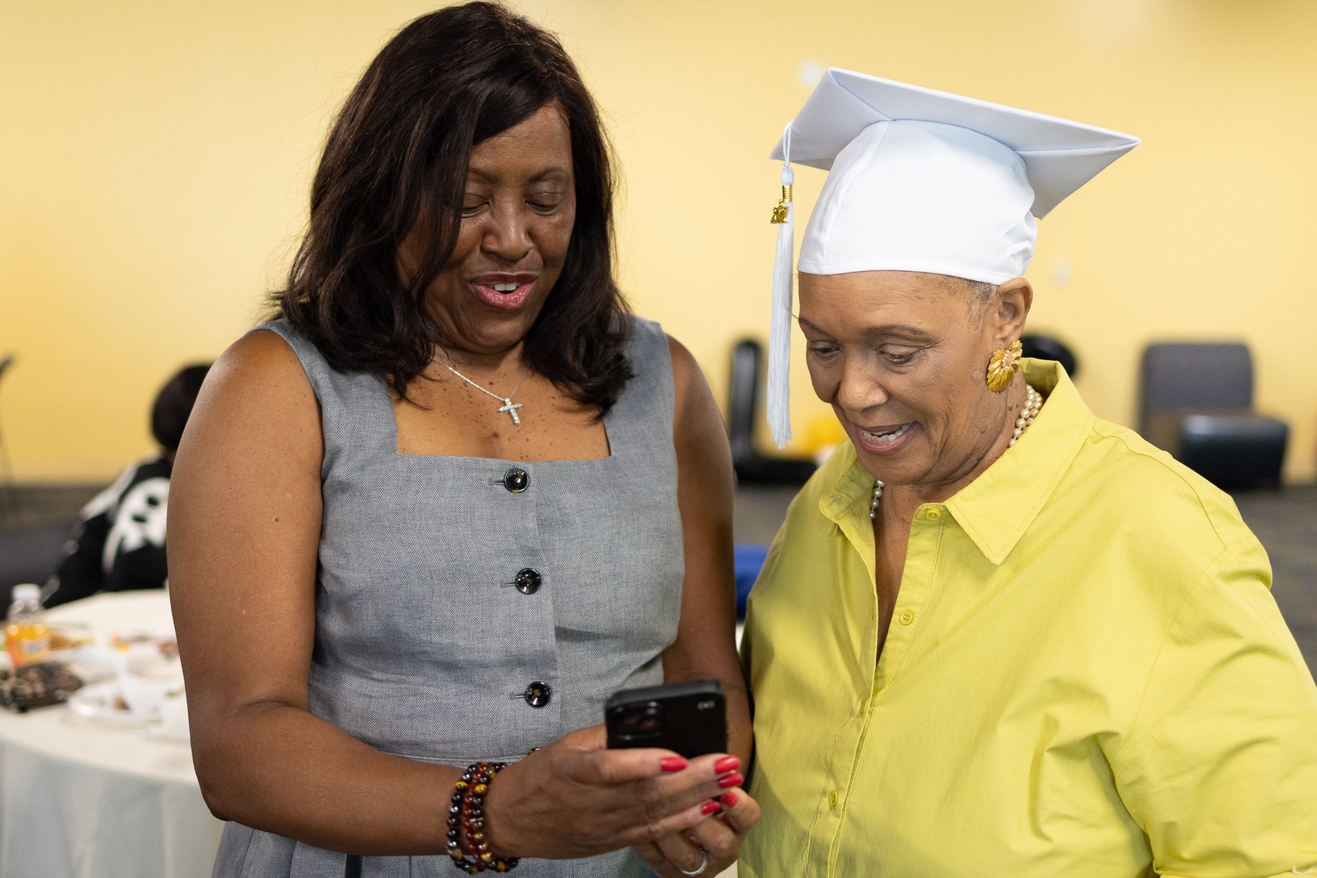 Woman showing cell phone to another woman wearing a graduation cap, both smiling, inside a building.