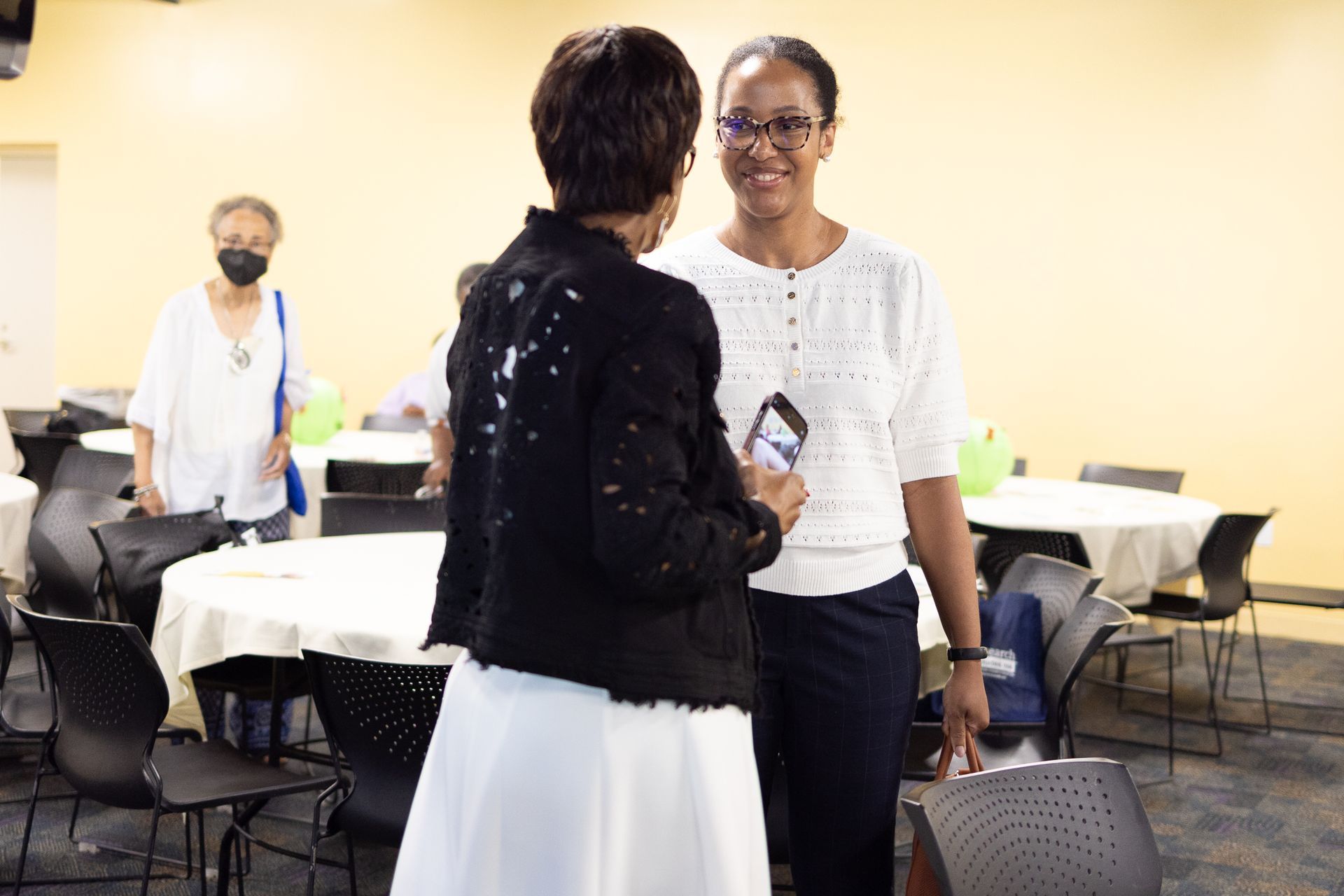 Two women conversing in a room with round tables and chairs. One holds a phone. A third person walks in the background.