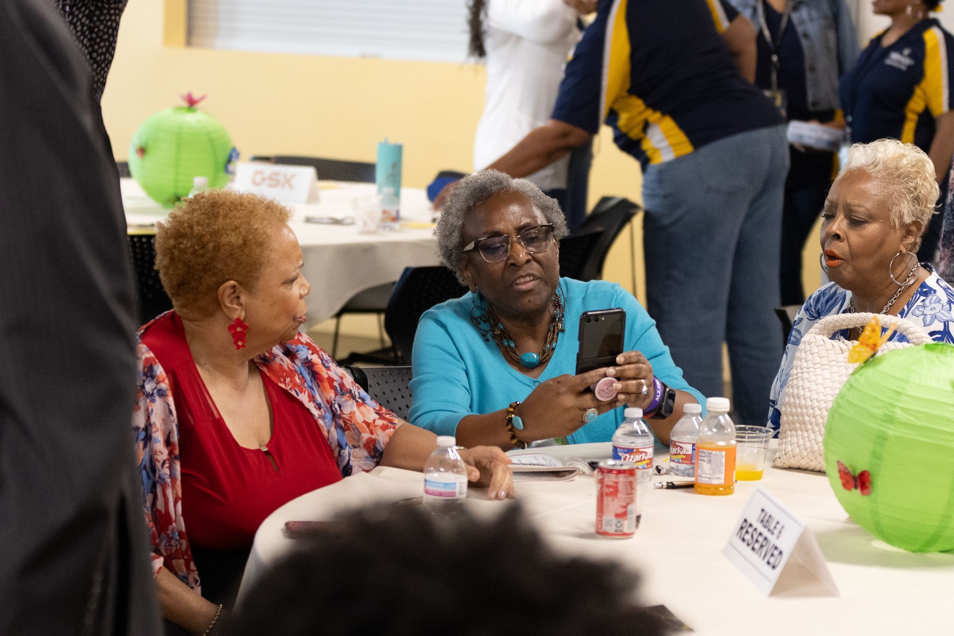 Three women seated at a table, two looking at the woman holding a phone. People are in the background.