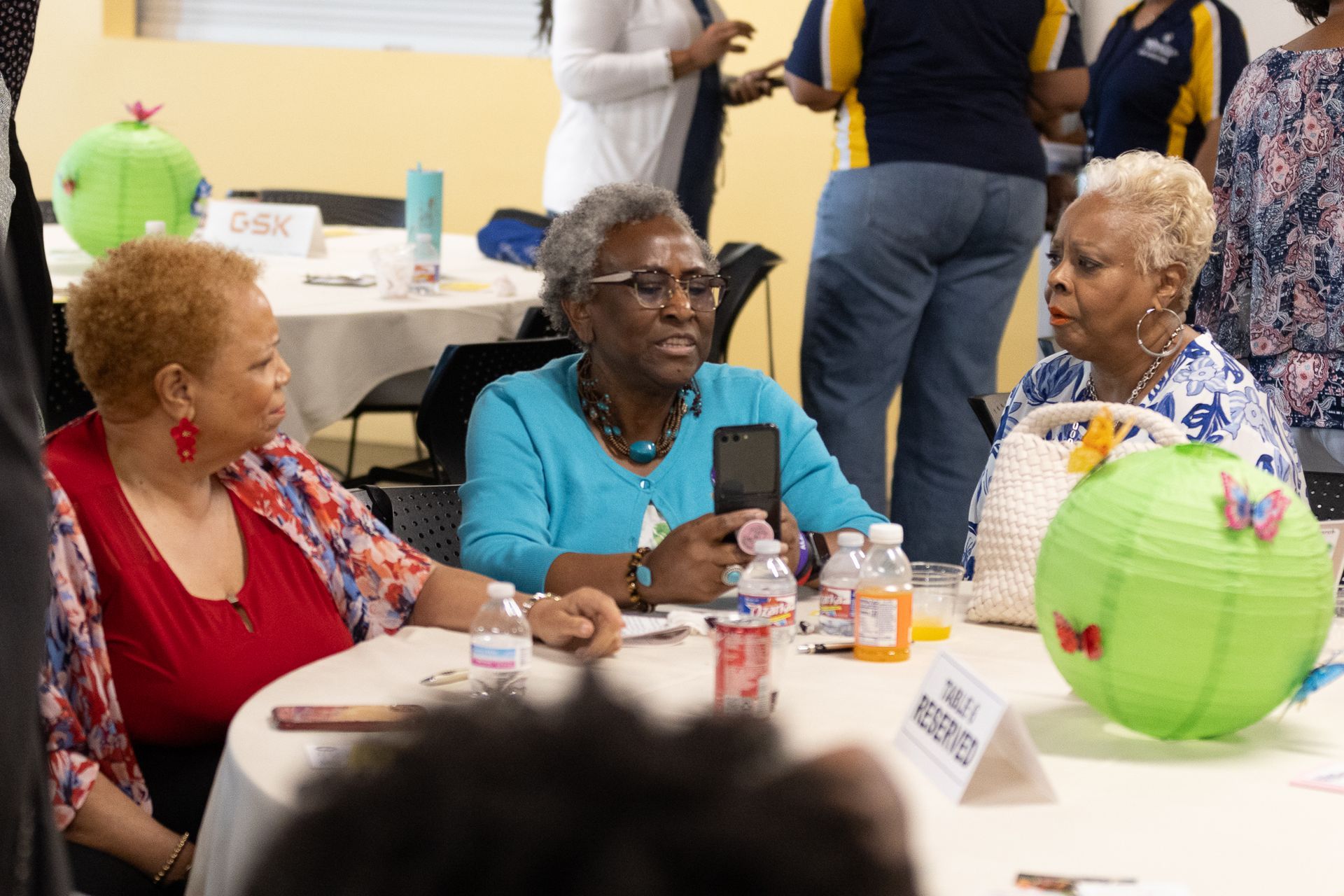 Three women at a table talking, one holding a phone. A green paper lantern is also on the table.