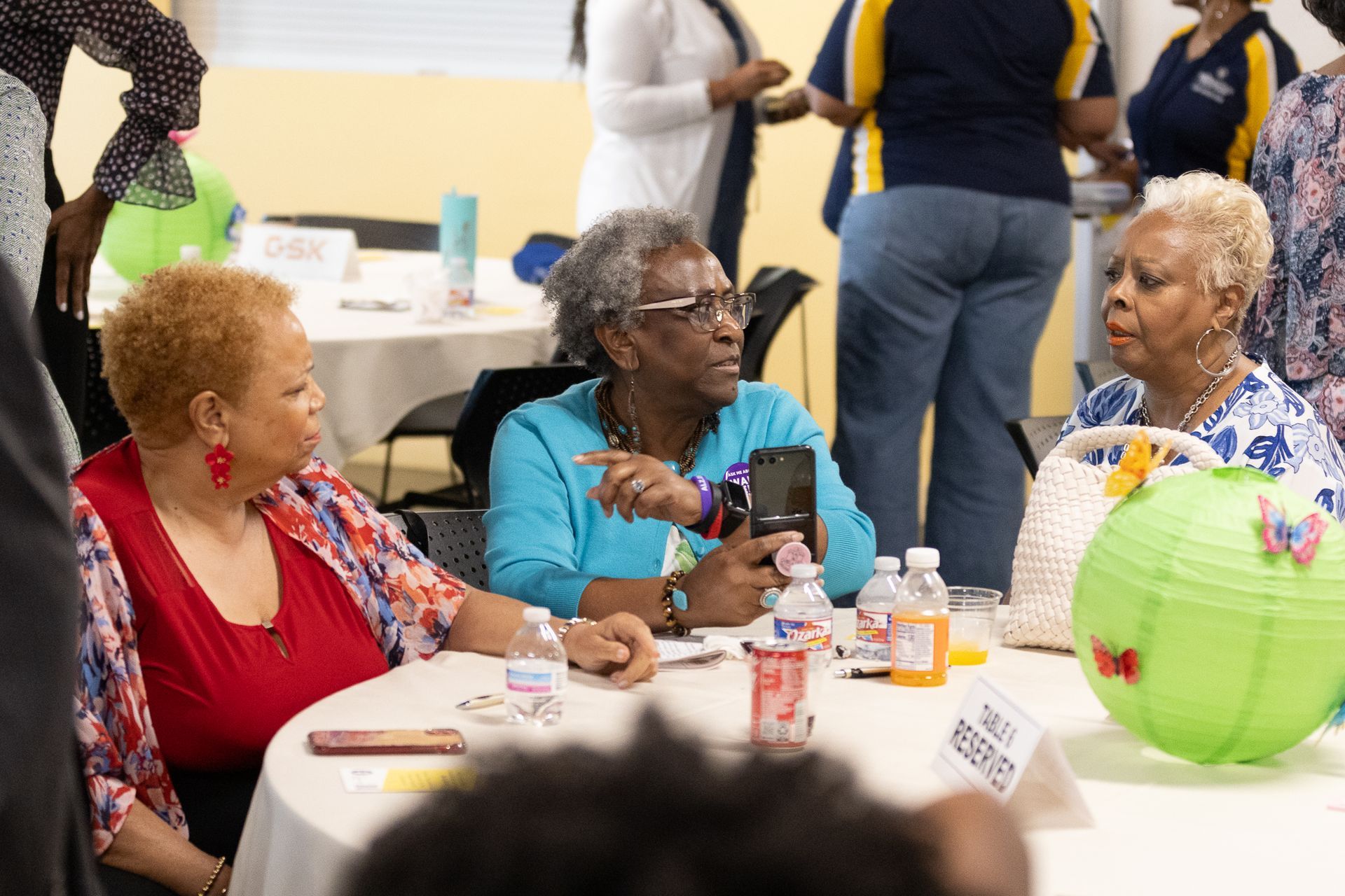 Three women seated at a table, conversing. One holds a phone. Setting appears to be an event or meeting.