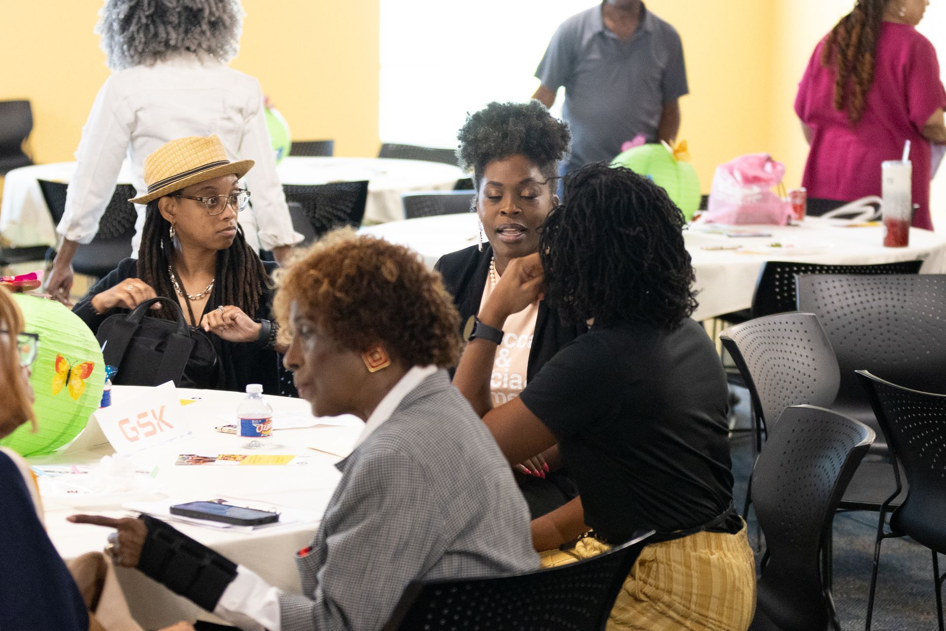 People seated at tables talking in a bright room. Some are holding papers or objects, looking at each other, and interacting.
