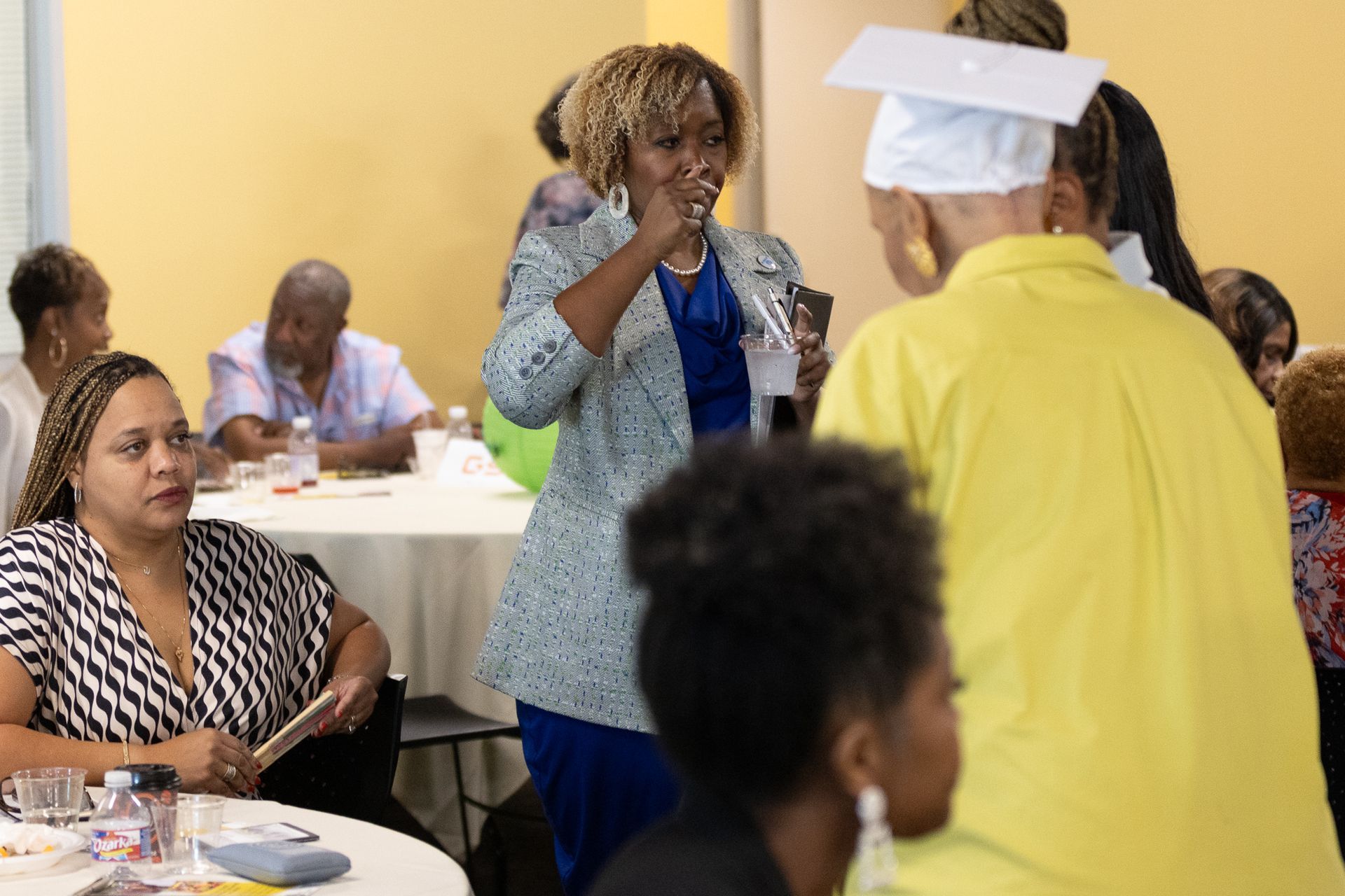 People at a gathering; woman in blazer speaks to person in yellow. Others sit at tables.