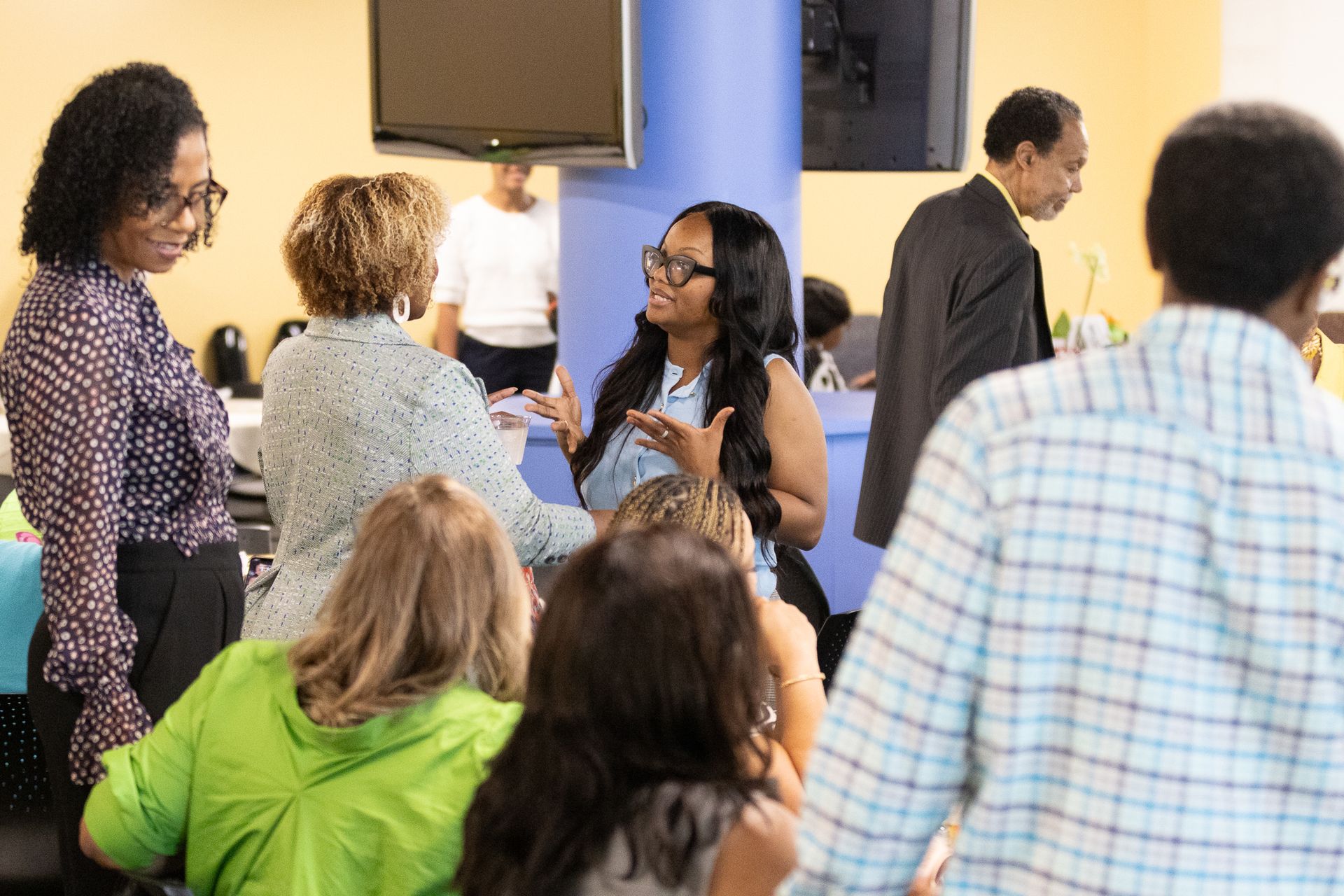 People in casual attire conversing in a brightly lit office setting. Some are sitting, others standing and gesturing.