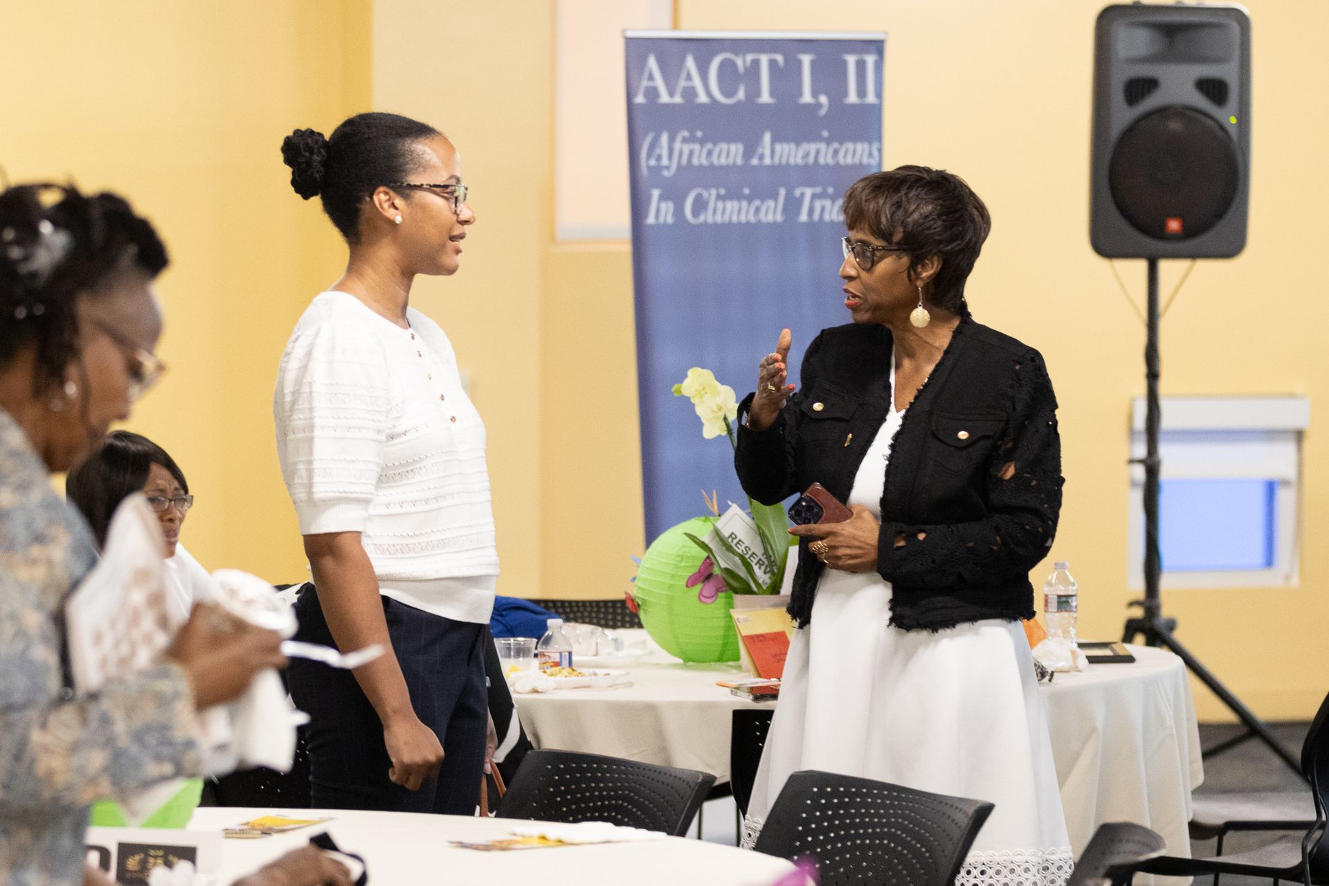 Two women converse at an event with a banner and tables. A woman gestures with her hand.