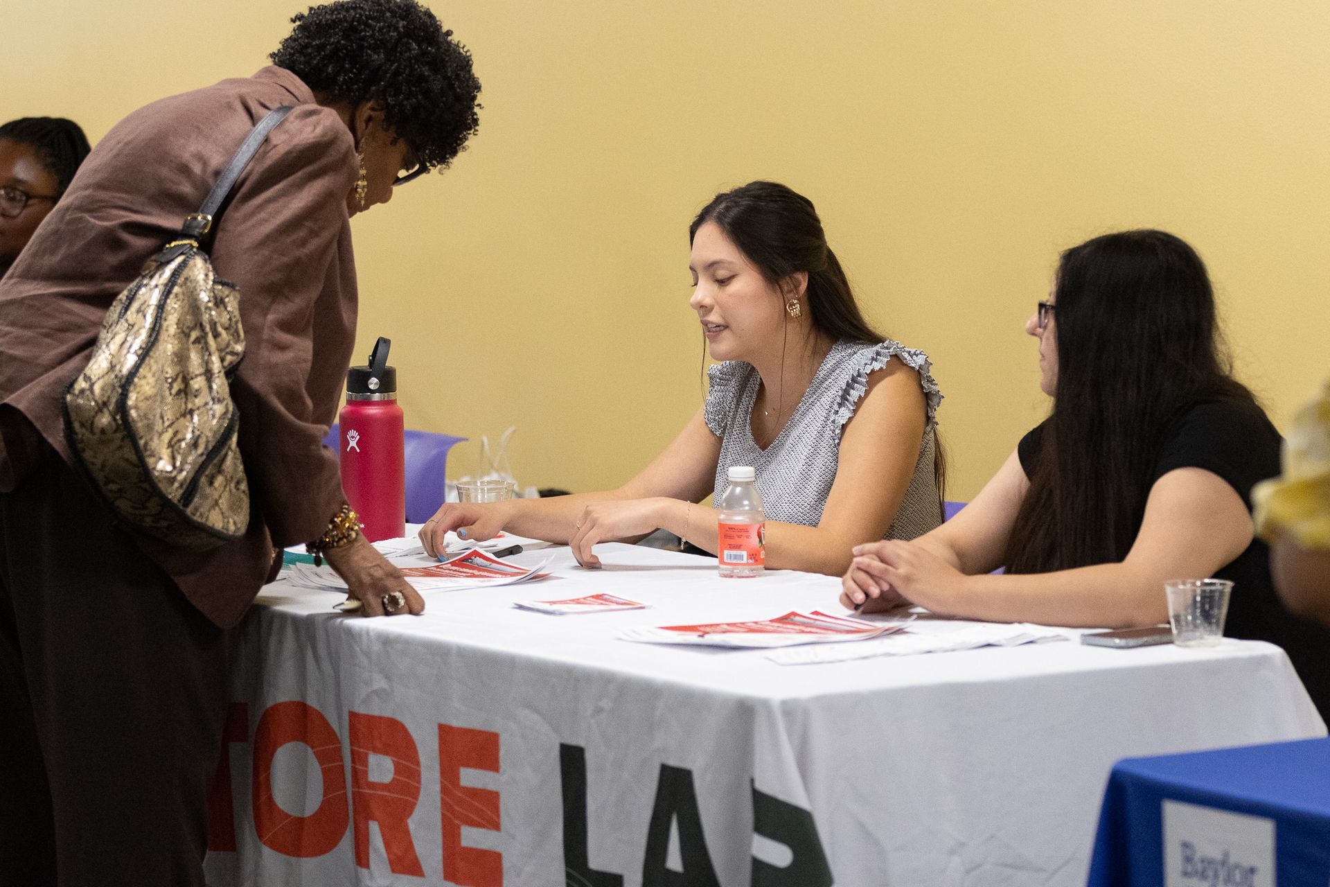 Three people at a table covered in a white cloth with red letters. One woman speaks to two others.