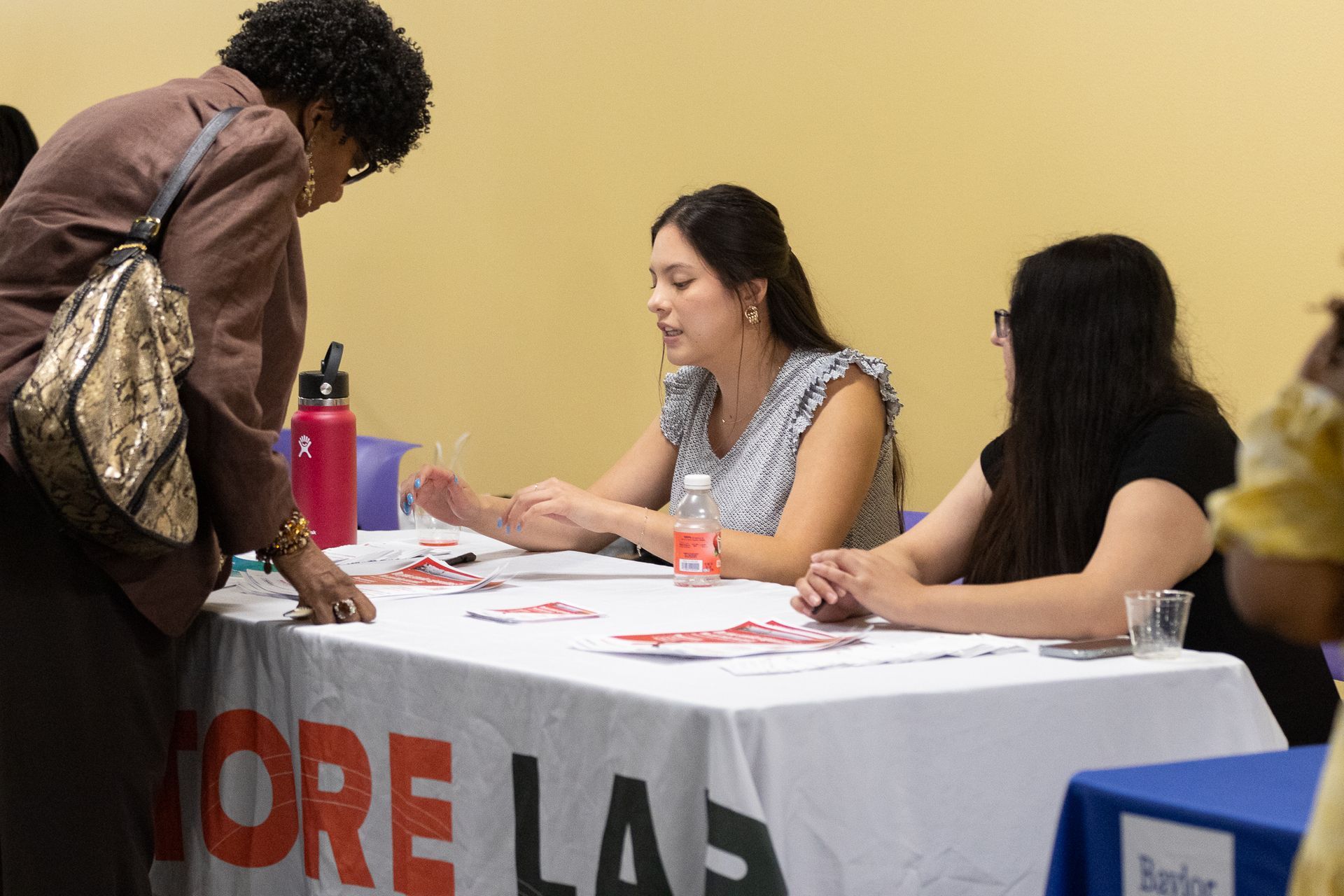 Woman talking at a table with two seated women. A banner on the table reads 