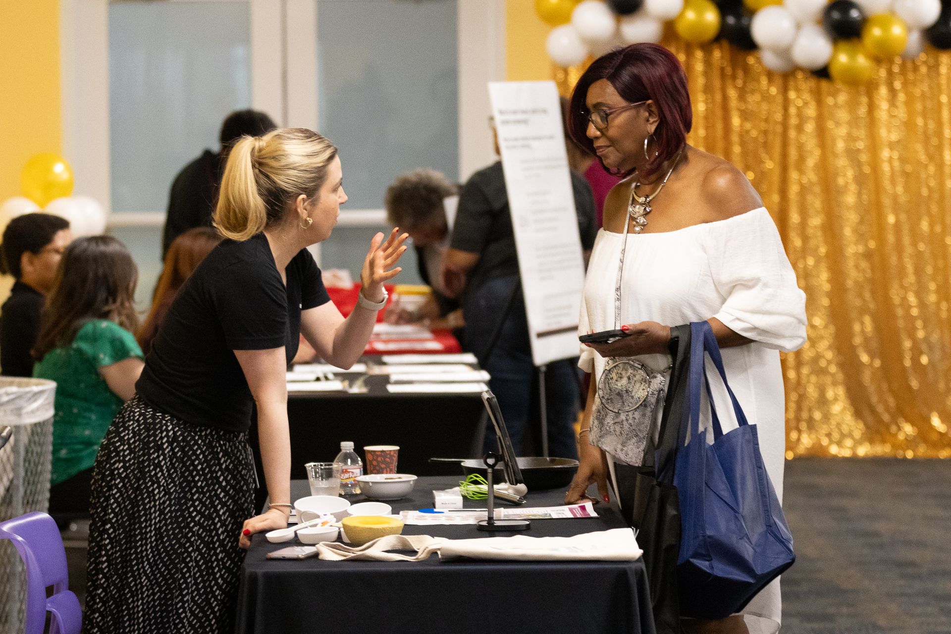 Two women at a table with information; one points, the other holds a phone. Event setting with balloons.