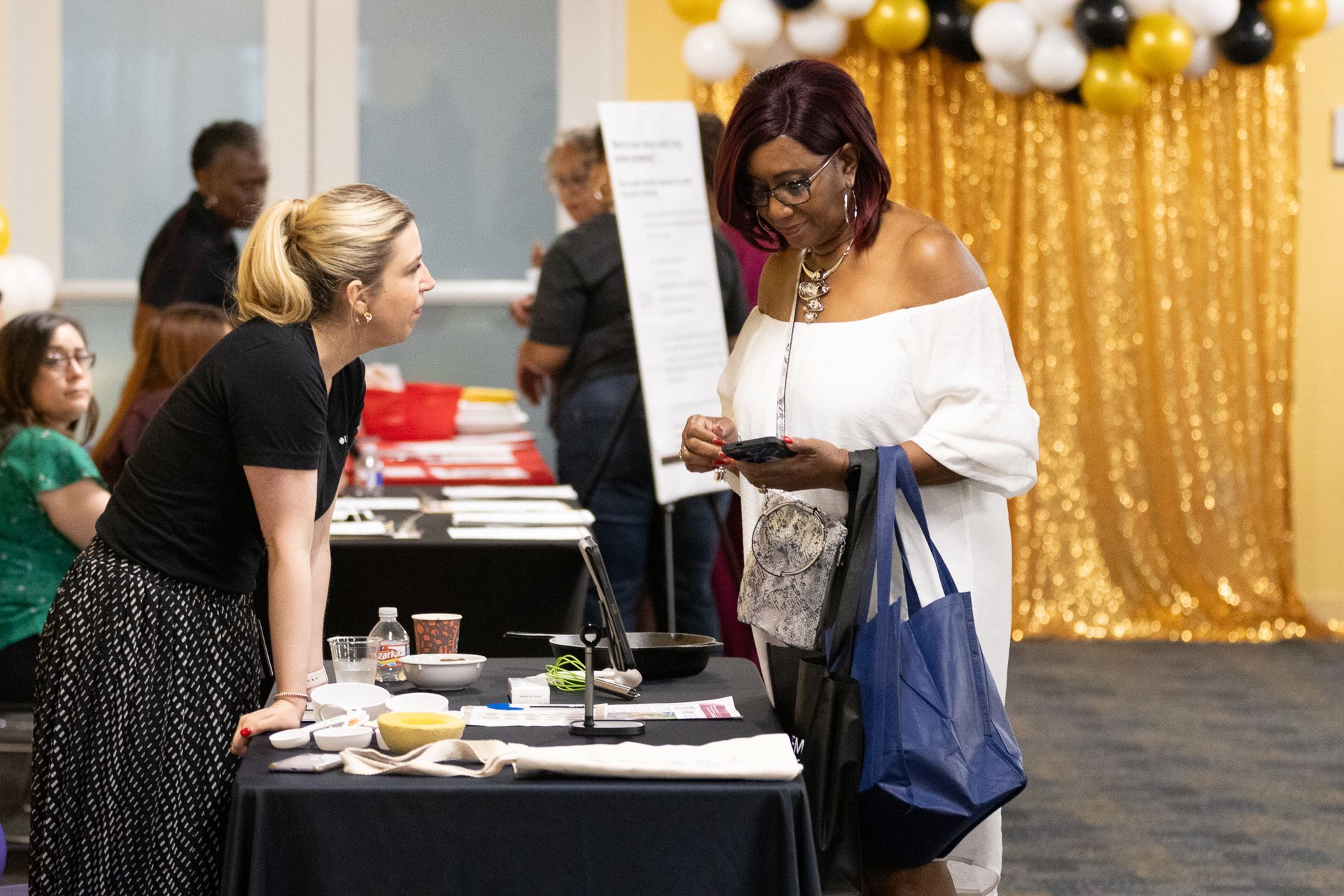 Woman in white dress looks at phone, talking with woman at a table with display. Gold curtain backdrop.