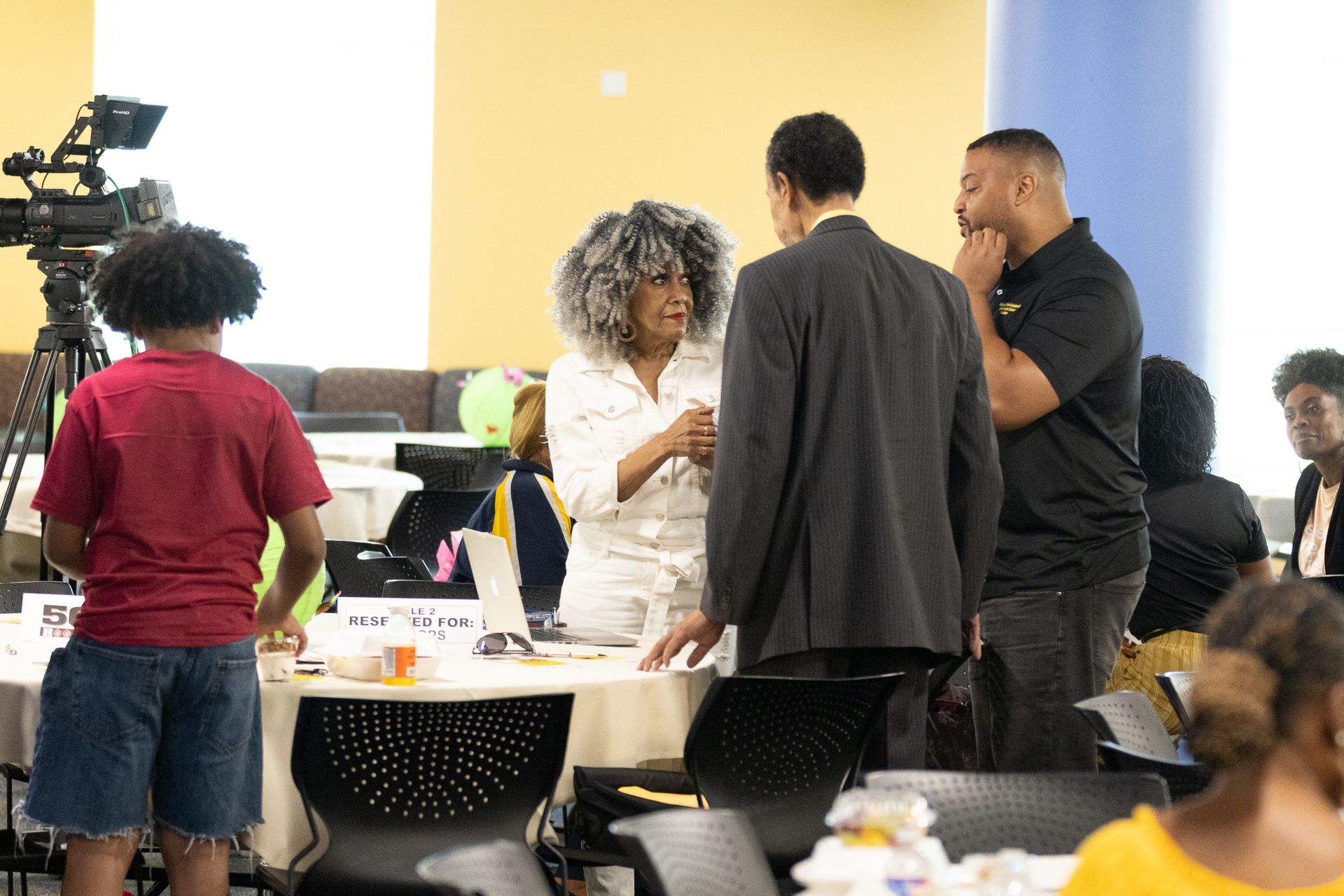 People in a room talking, seated at tables. A camera on a tripod is on the left.