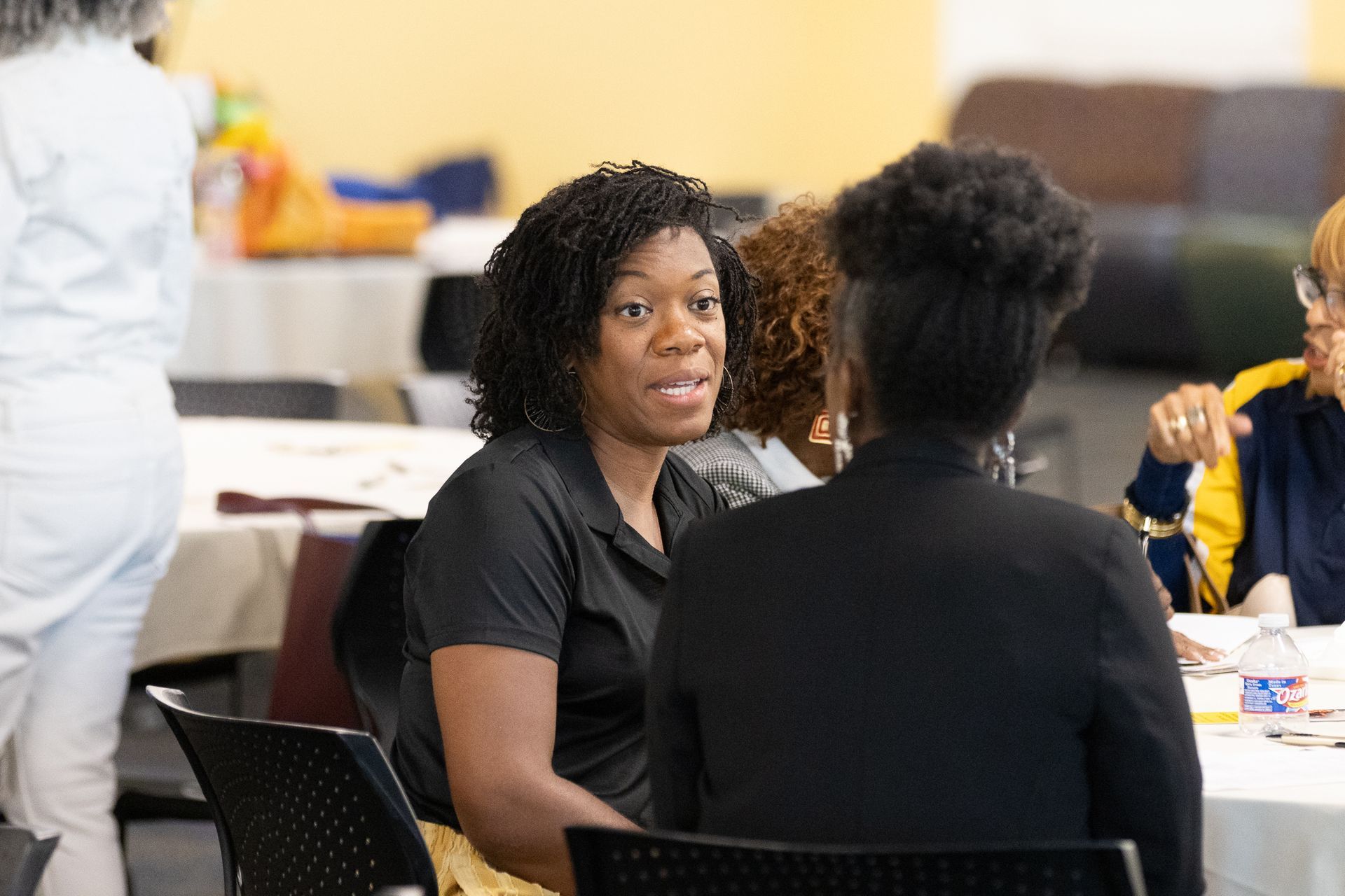 Woman in black shirt speaks to another woman at a table, indoor setting.