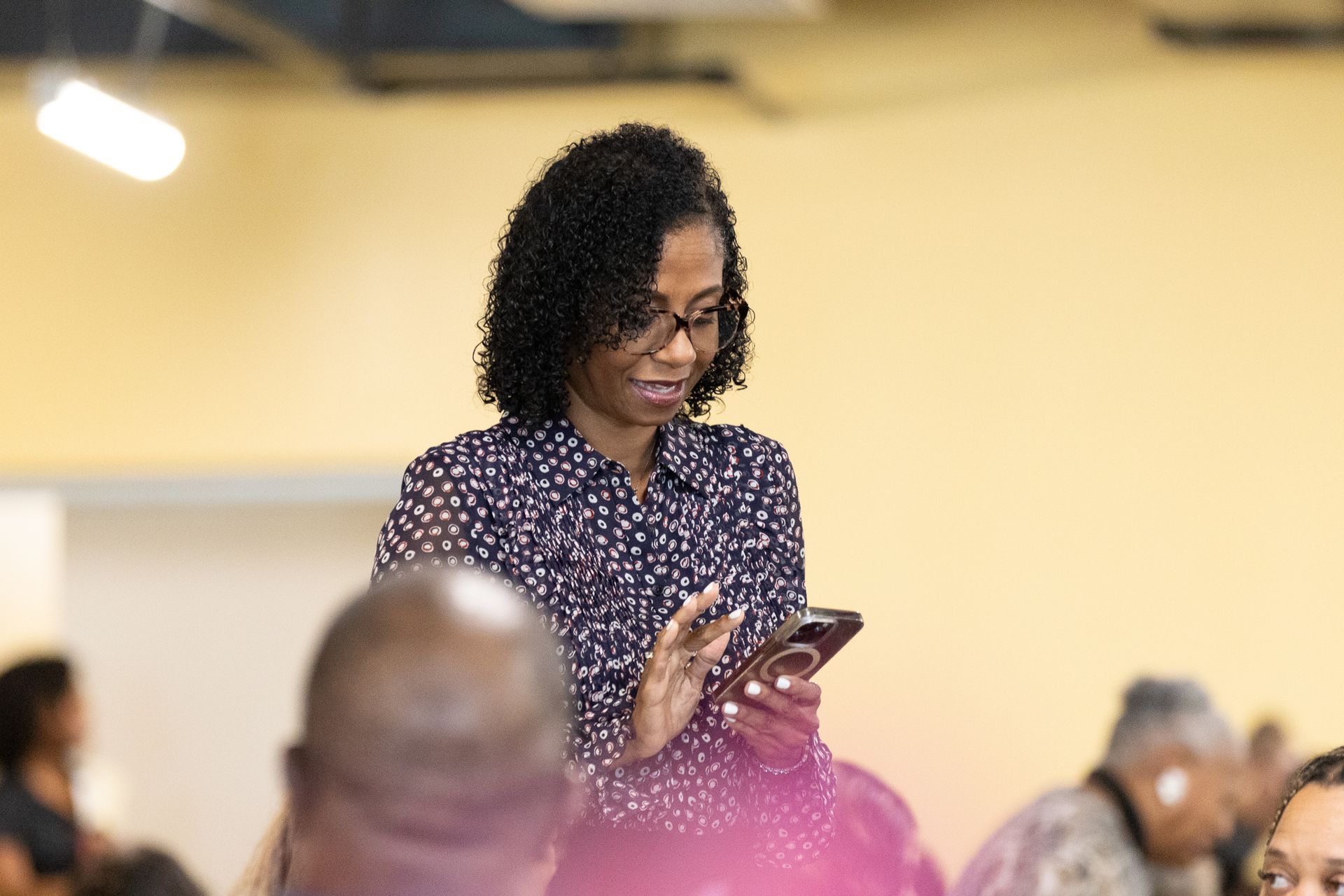 Woman with glasses and patterned shirt looks at a phone in a room with a crowd.