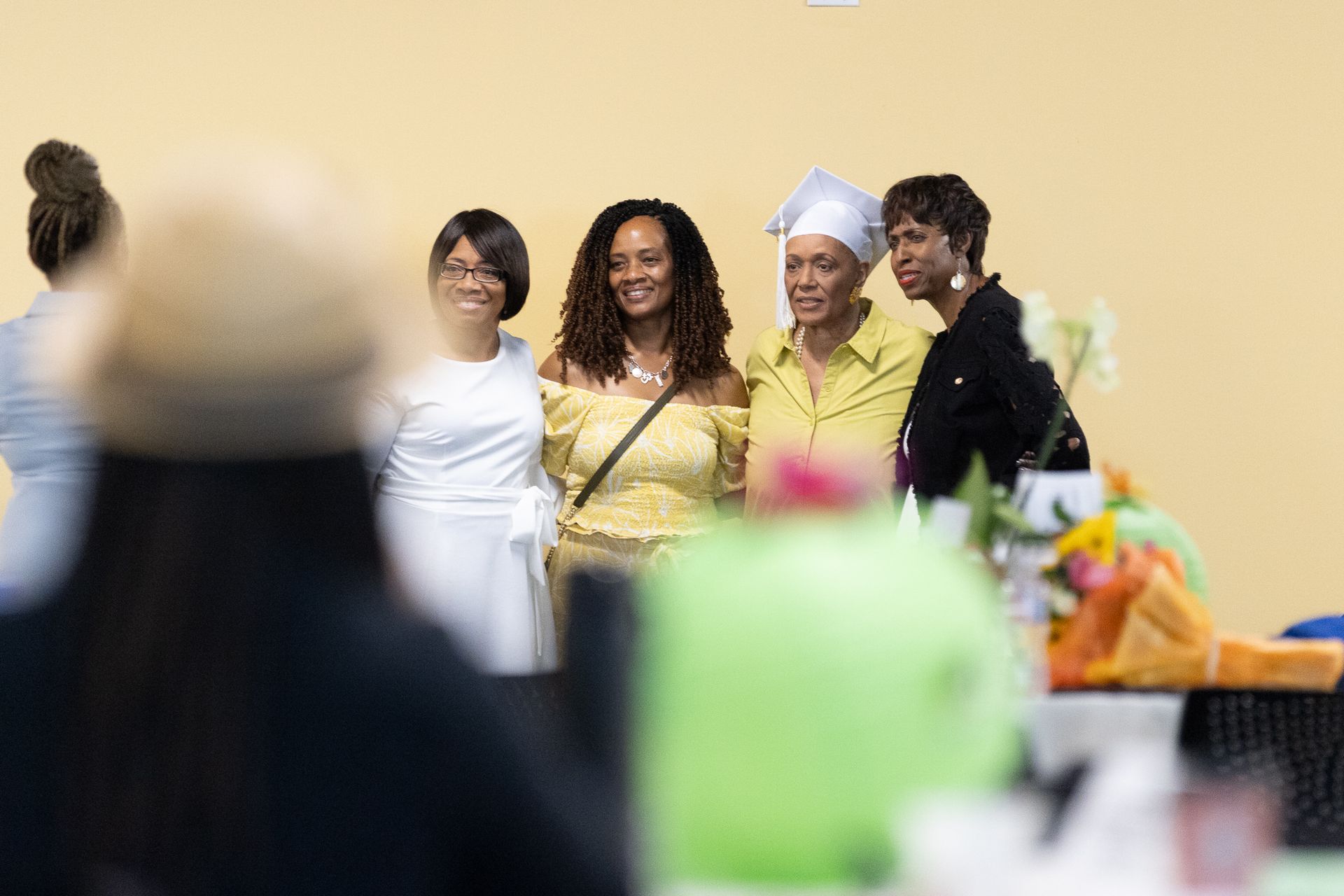 Four women smiling, posing together indoors. A table with gifts is in the foreground.