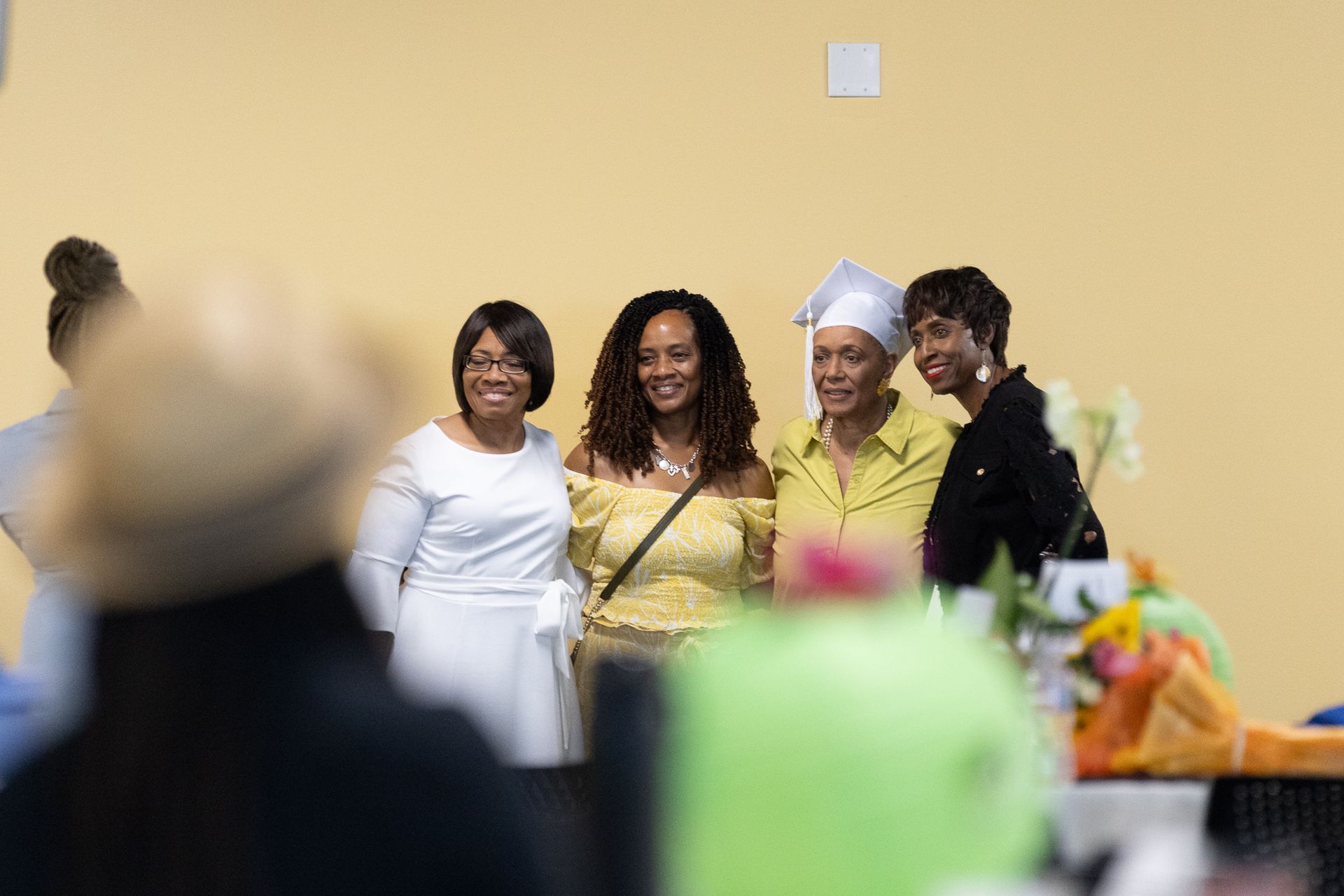 Four smiling people pose for a photo in front of a tan wall. One wears a graduation cap.
