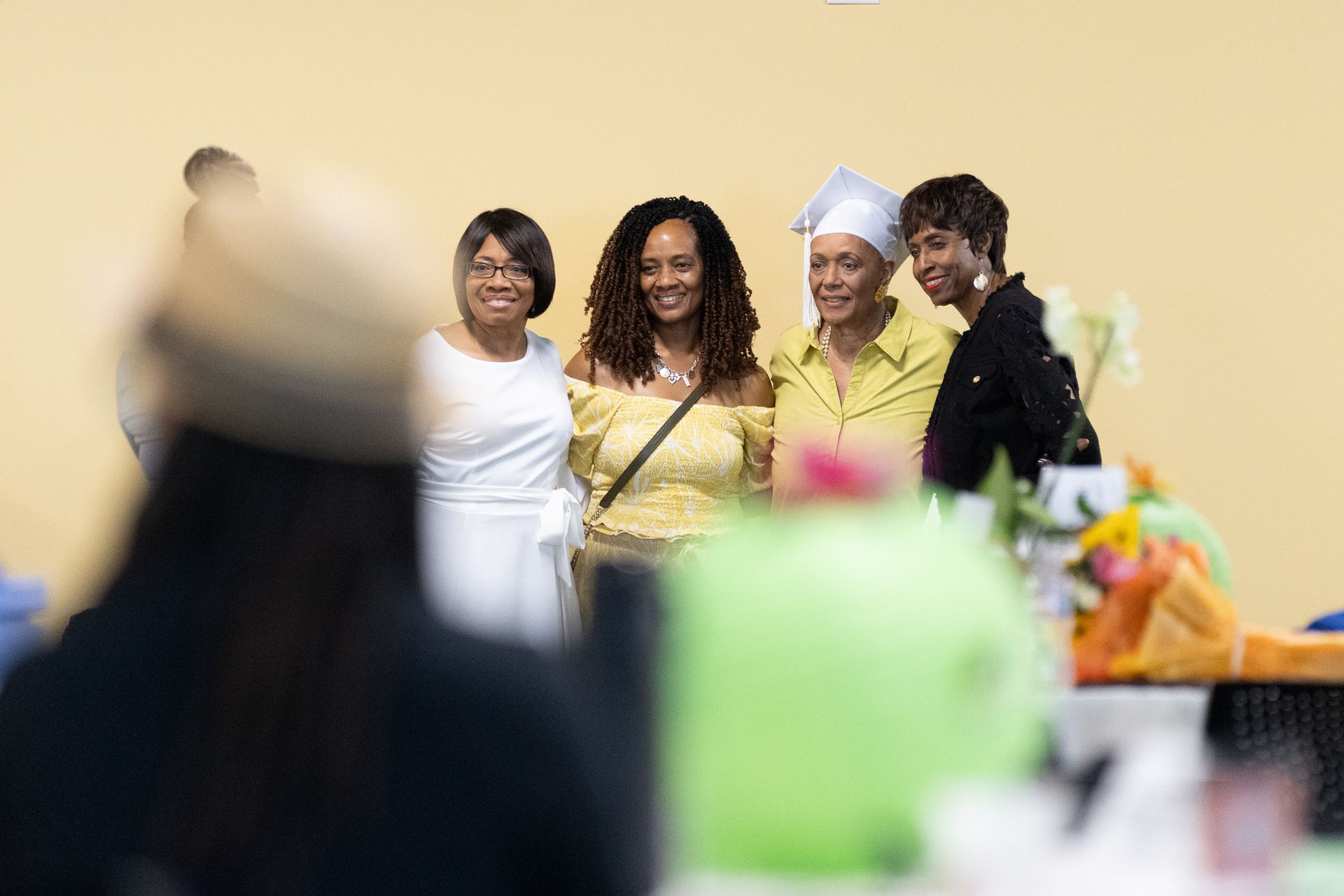 Four smiling women posing for a photo at a graduation ceremony. One wears a cap and gown.